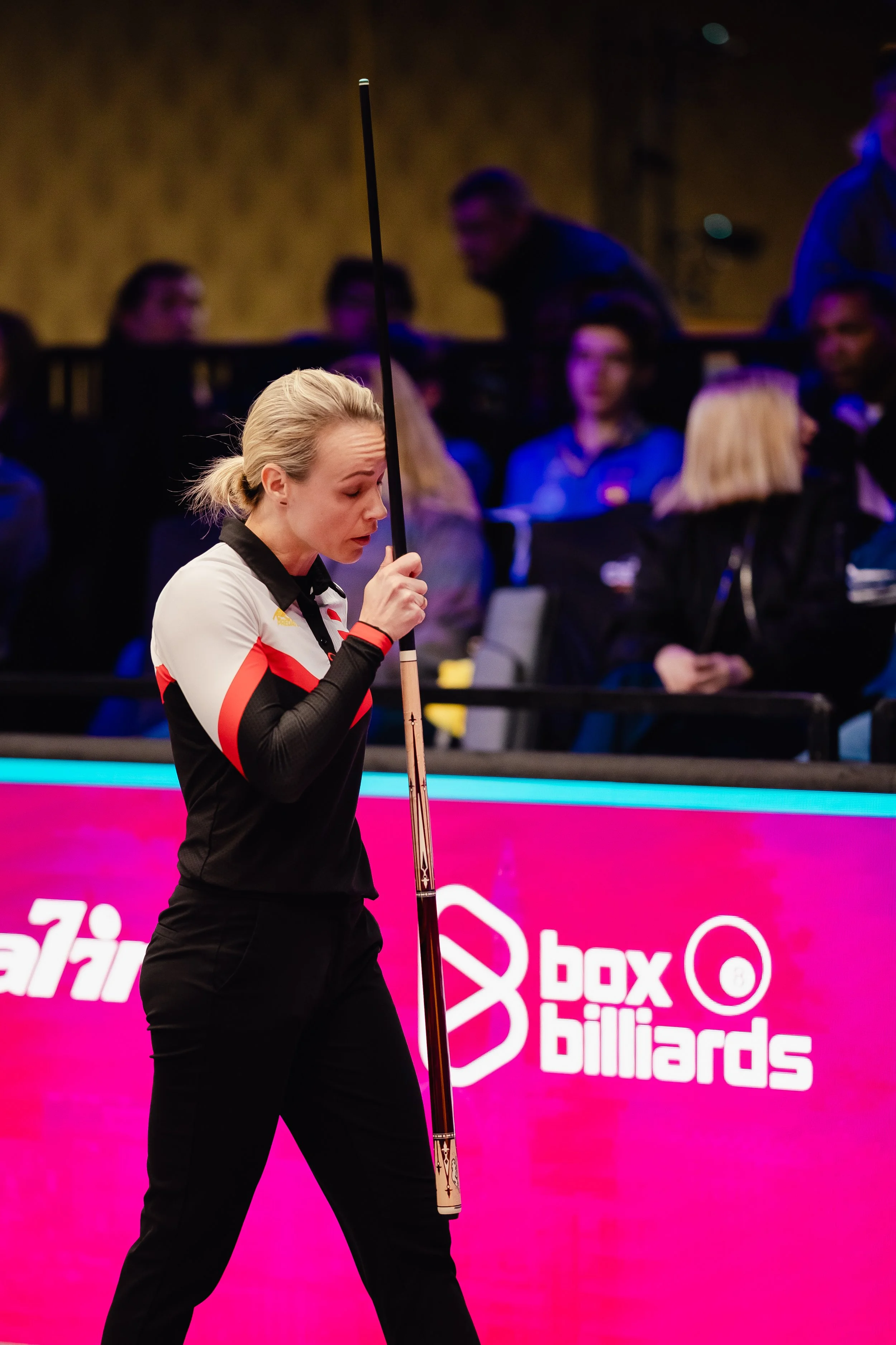 A female billiards player holding a cue stick during a game at an indoor billiards venue, with an audience in the background and a pink digital display board that says 'box billiards'.