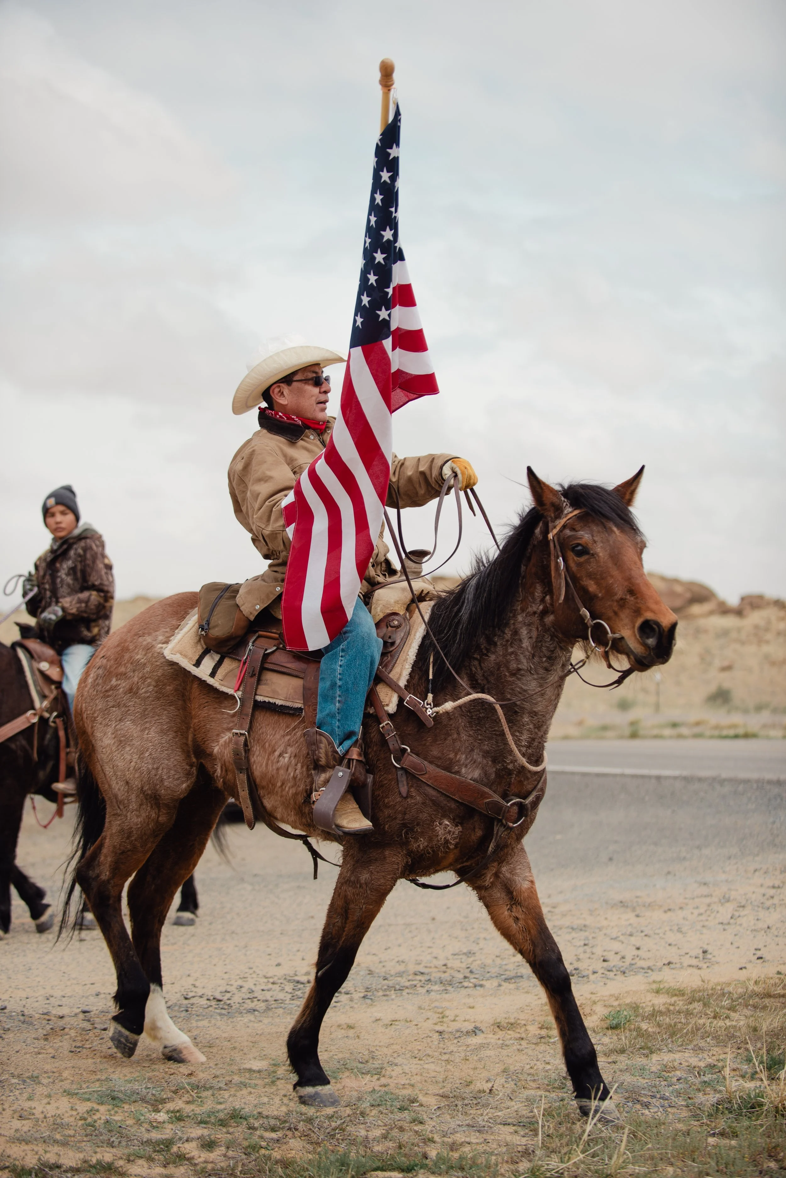 A man riding a horse, holding an American flag, with a boy in the background riding another horse in a desert landscape under a cloudy sky.