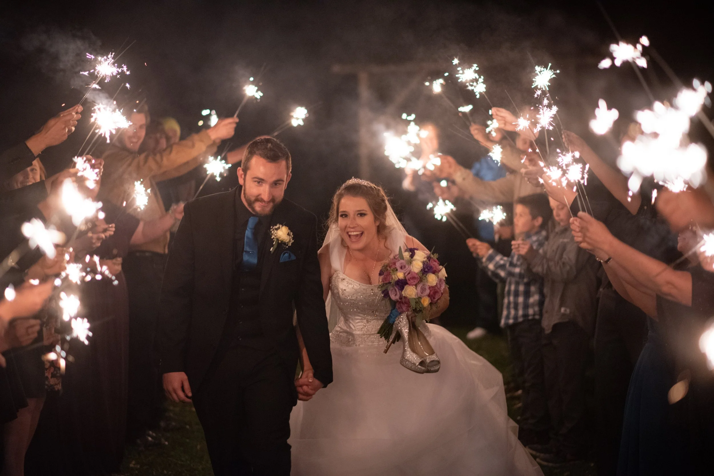 Newlywed couple holding hands and smiling, surrounded by guests holding sparklers at night.