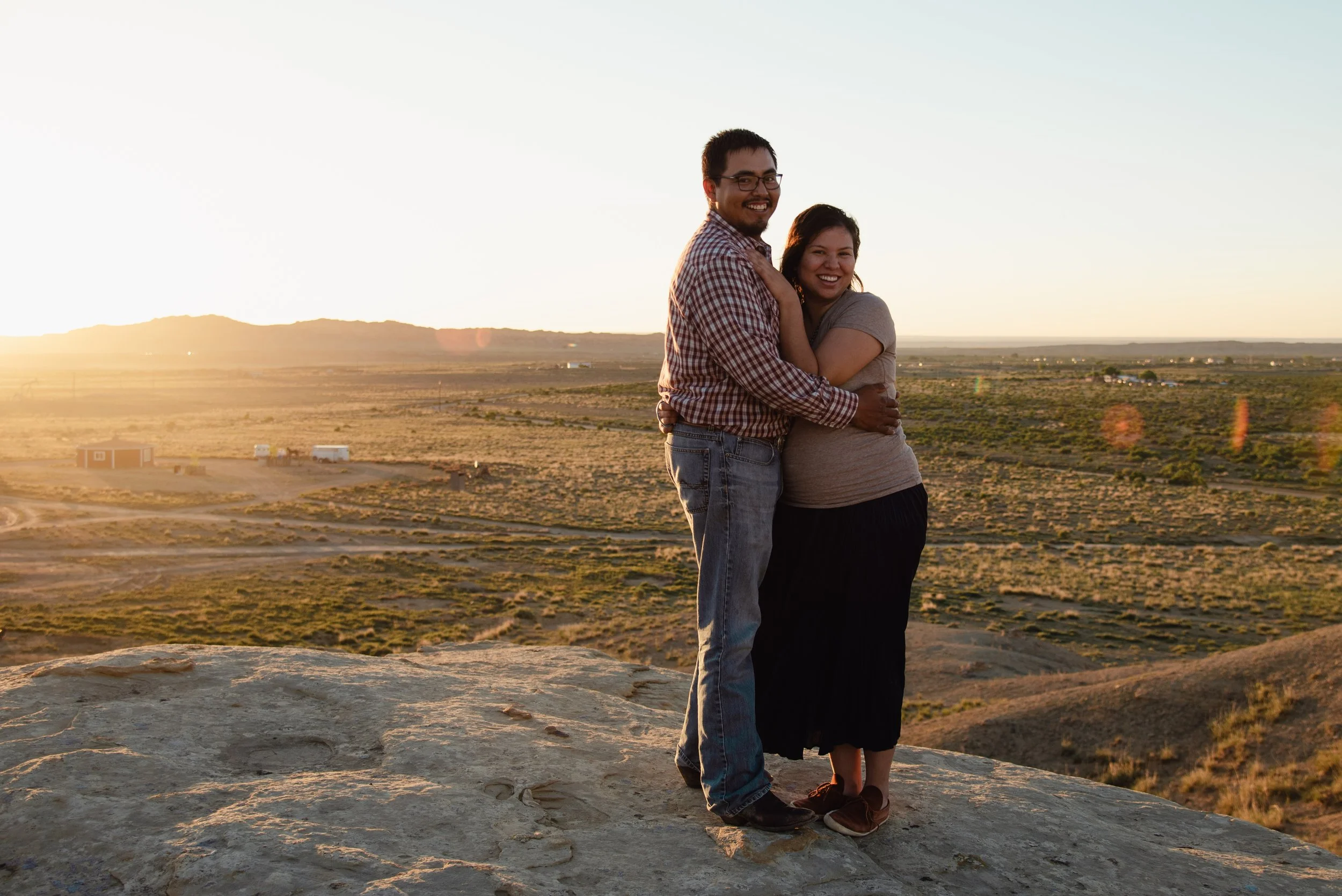 A happy couple standing on a rock at sunset, embracing, with a vast open plain and distant hills in the background.