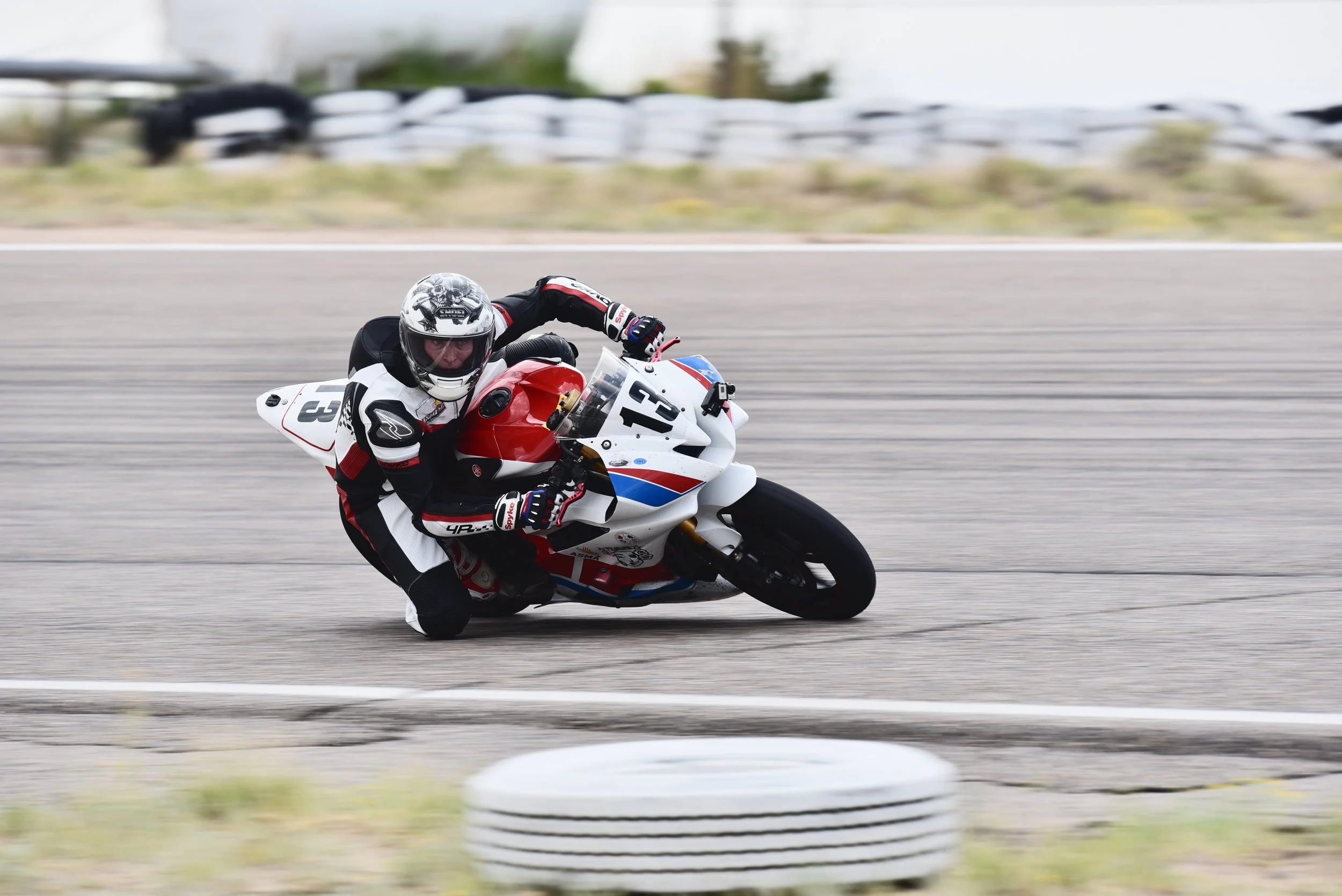 Motorcycle racer leaning into a turn on a racetrack, wearing a black and white racing suit and helmet, with the motorcycle numbered 13, red, white, and blue color scheme.