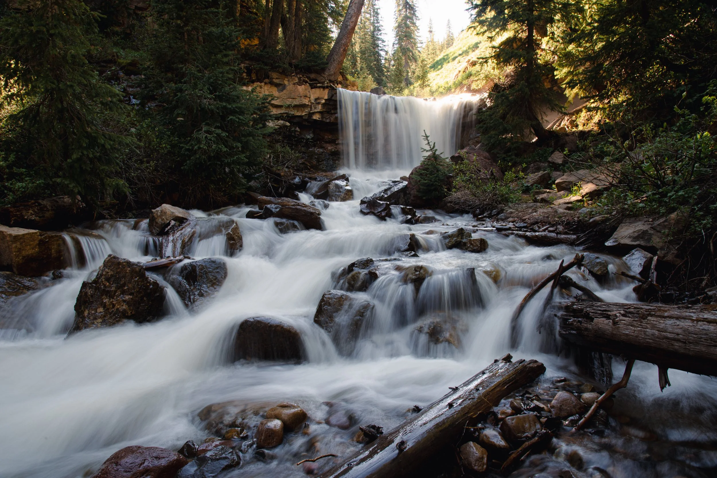 A serene mountain stream flowing over rocks with a small waterfall in a forested area, sunlit trees in the background.