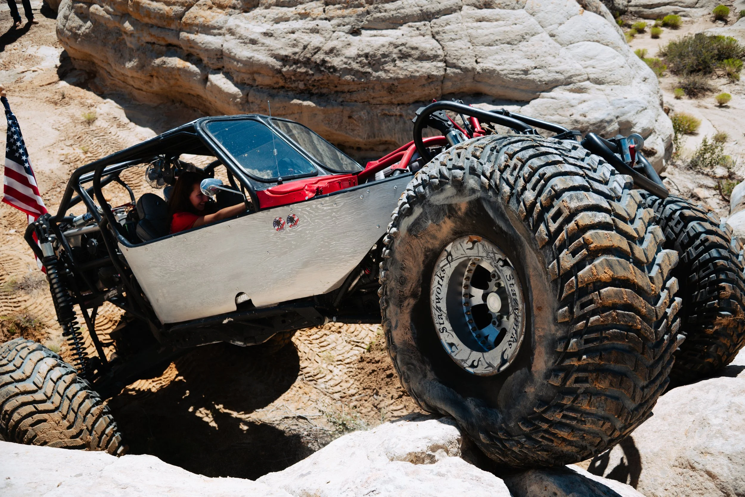 A custom off-road vehicle with large tires navigating sandy, rocky terrain, driven by a woman inside.