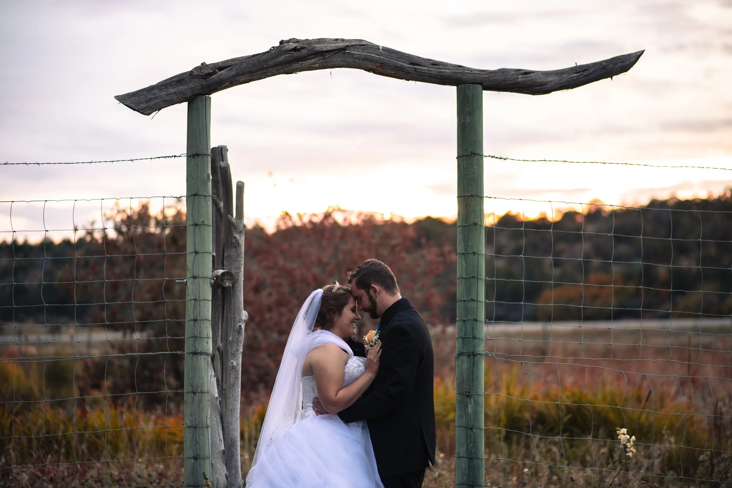 A bride and groom standing close together under a wooden arch in an outdoor setting during sunset, embracing affectionately.