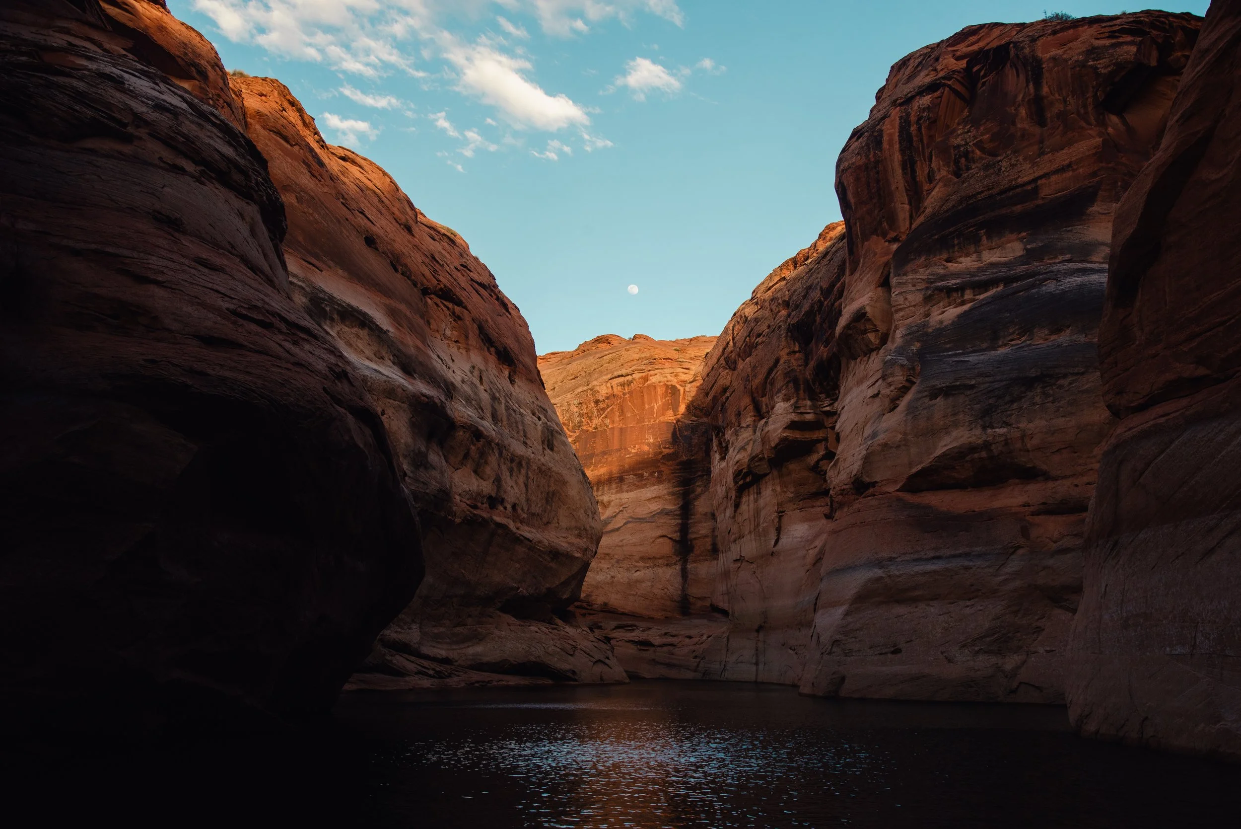 A canyon with tall, reddish-brown rock walls and a body of water at the bottom, under a clear blue sky with a few clouds and the moon visible.
