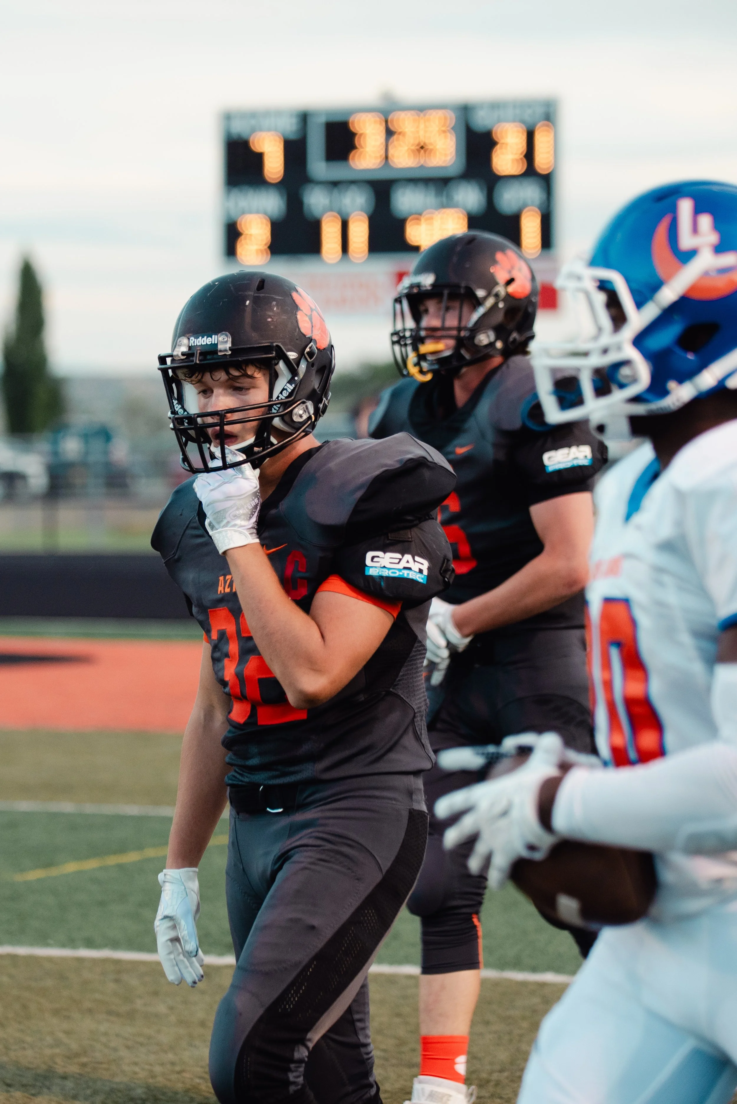 American football players on the field before a game or during a break, with a digital scoreboard in the background.