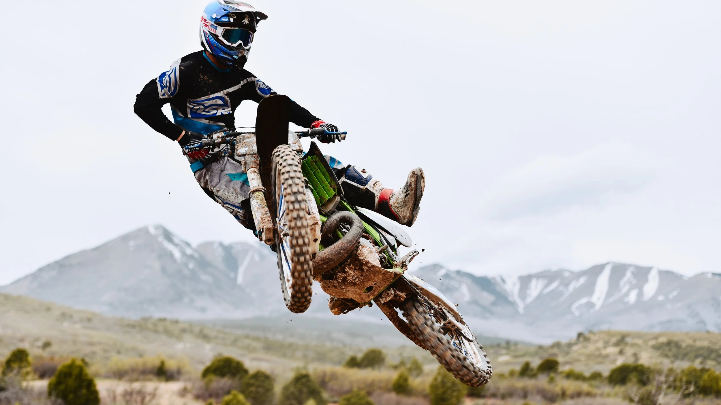 Motocross rider mid-air on a dirt bike, performing a jump over rugged terrain with mountains in the background.