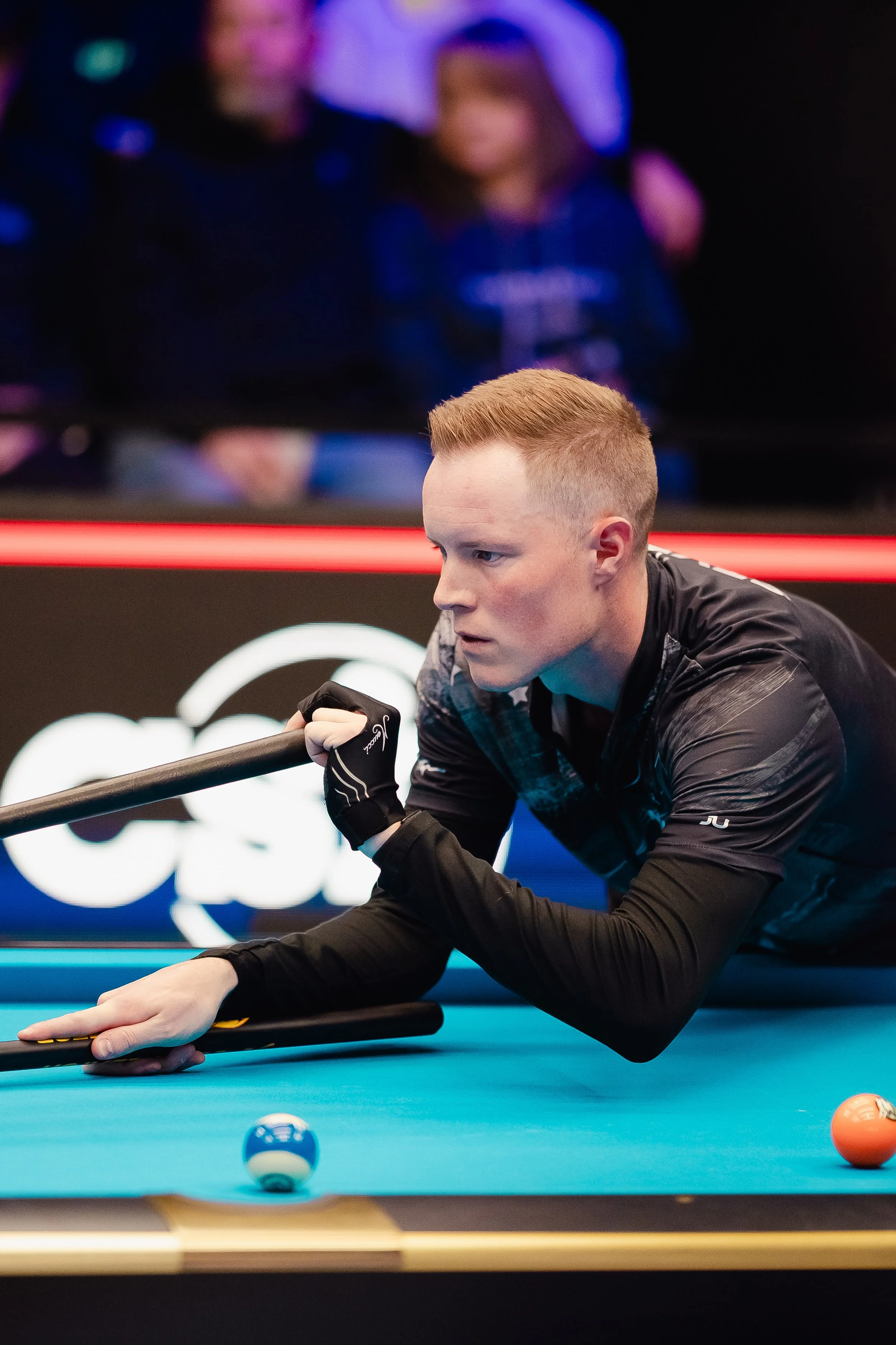 A young man with short red hair playing pool, focused on making a shot, wearing a black long-sleeve shirt and gloves, on a bright blue pool table with colored balls.