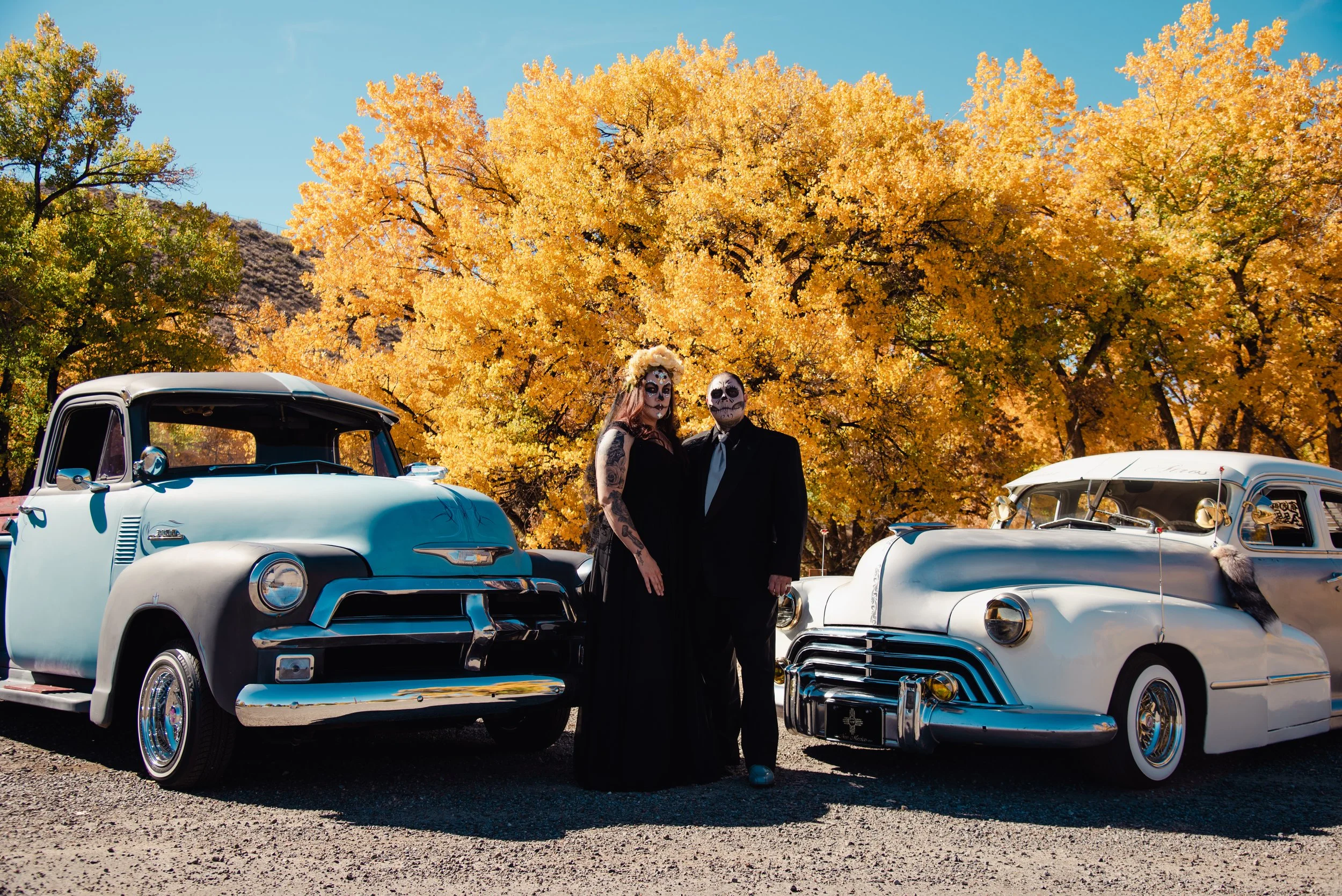 Couple with Day of the Dead face paint standing between vintage cars in front of autumn-colored trees.