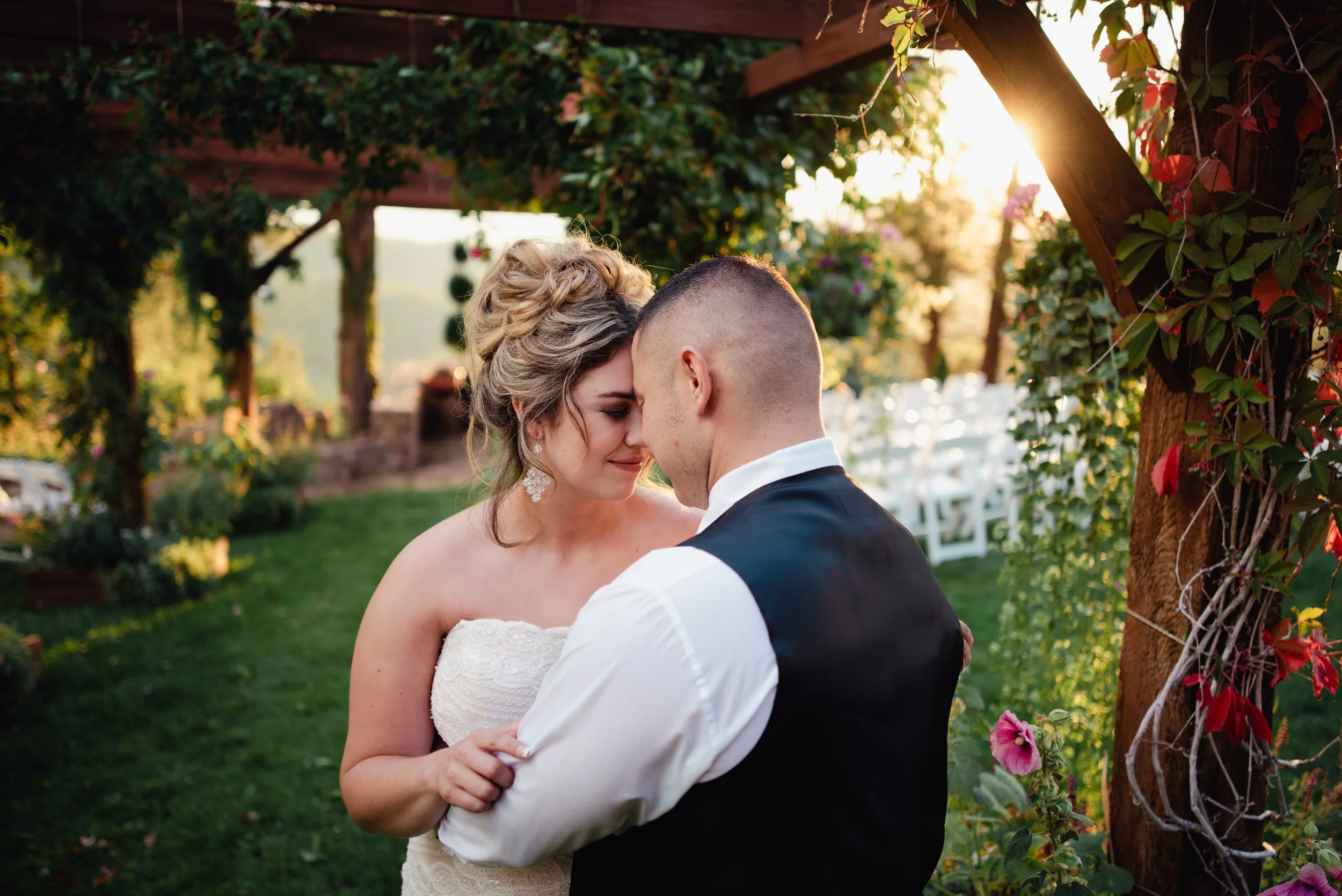 A bride and groom sharing a romantic moment at sunset in a garden with greenery and flowers.