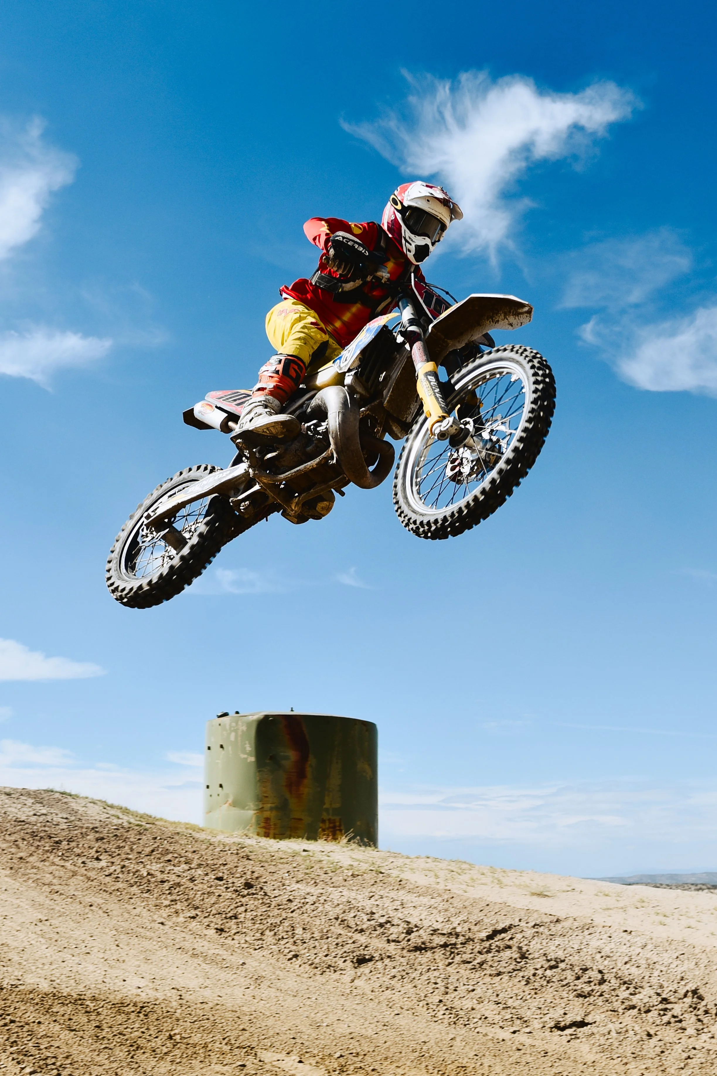Motorcyclist in red and yellow gear jumping over a metal barrel on a dirt track under blue sky.