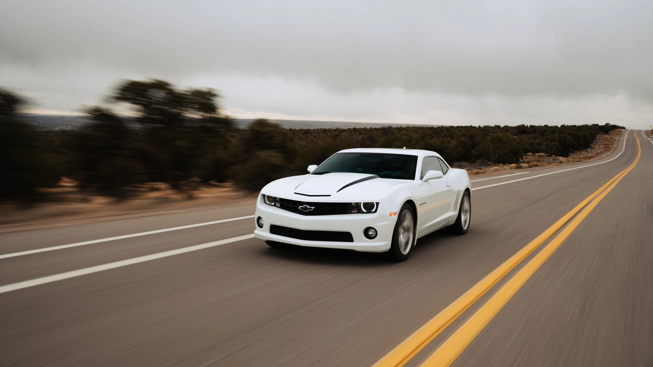 A white Chevrolet Camaro sports car driving on a winding two-lane road through a semi-arid landscape under overcast skies.