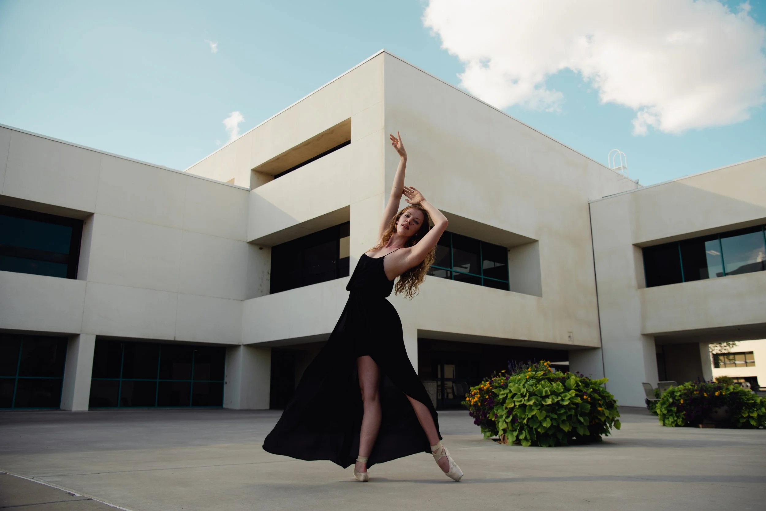 A woman in a black dress dancing ballet outdoors in front of a modern white building with flower pots.