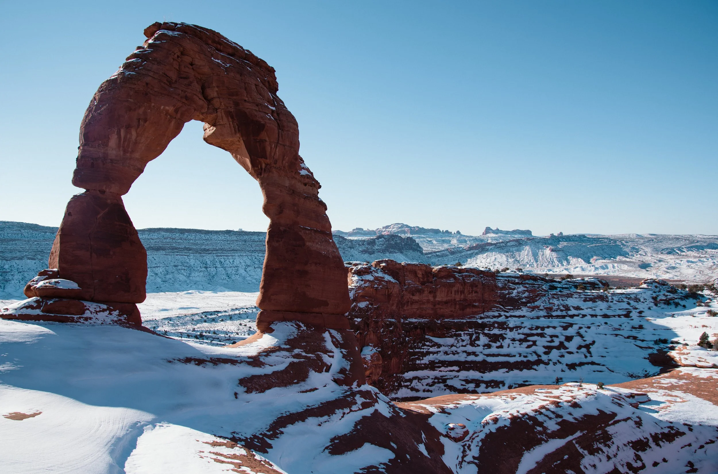 Snow-covered desert landscape featuring the iconic Delicate Arch in Arches National Park, Utah, with clear blue sky in the background.