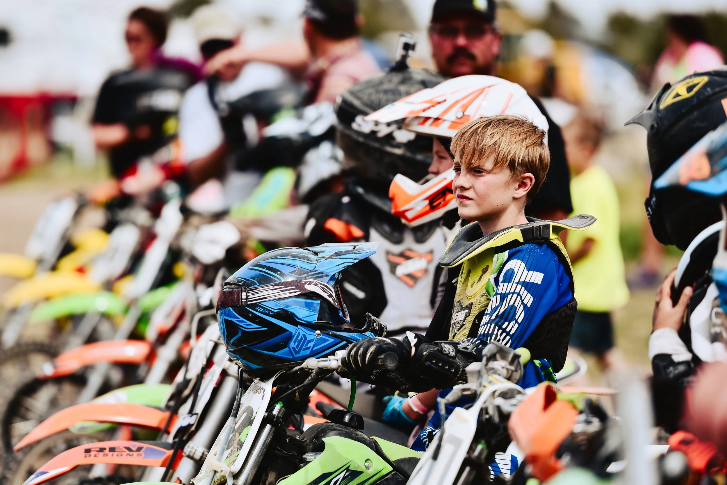 Young boy in motocross gear sitting on a dirt bike surrounded by other riders and bikes, with spectators in the background.