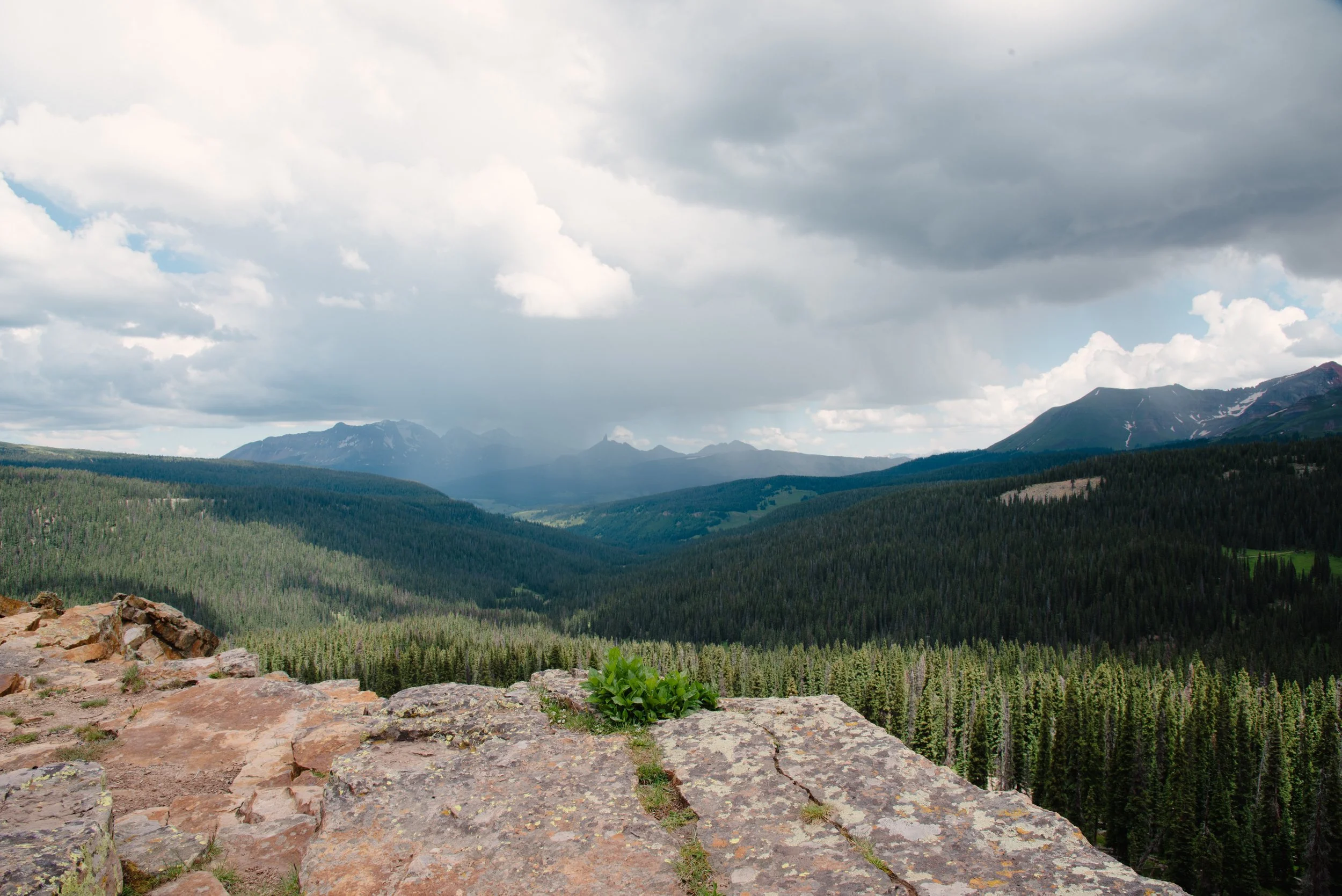 Scenic view of a valley with dense evergreen forests, mountains in the distance, and clouds overhead, as seen from a rocky overlook.