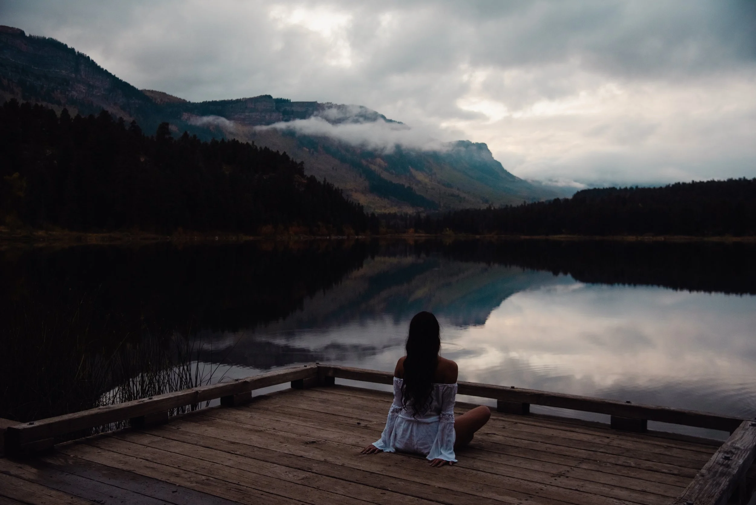 A woman sitting on a wooden dock, facing a calm lake and mountains under a cloudy sky.