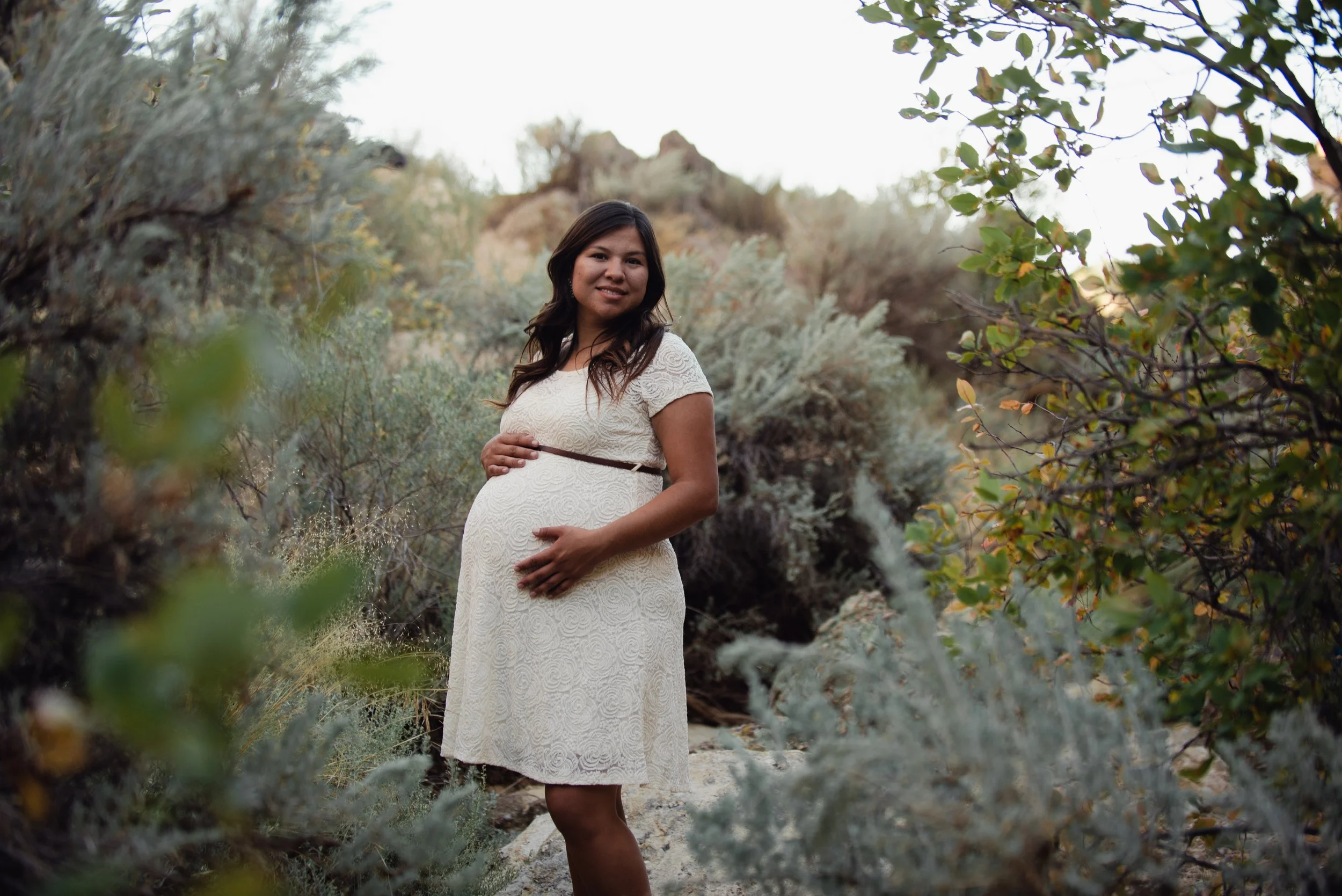 A pregnant woman standing in a natural, desert-like landscape with bushes and rocks, wearing a white lace dress and smiling at the camera.