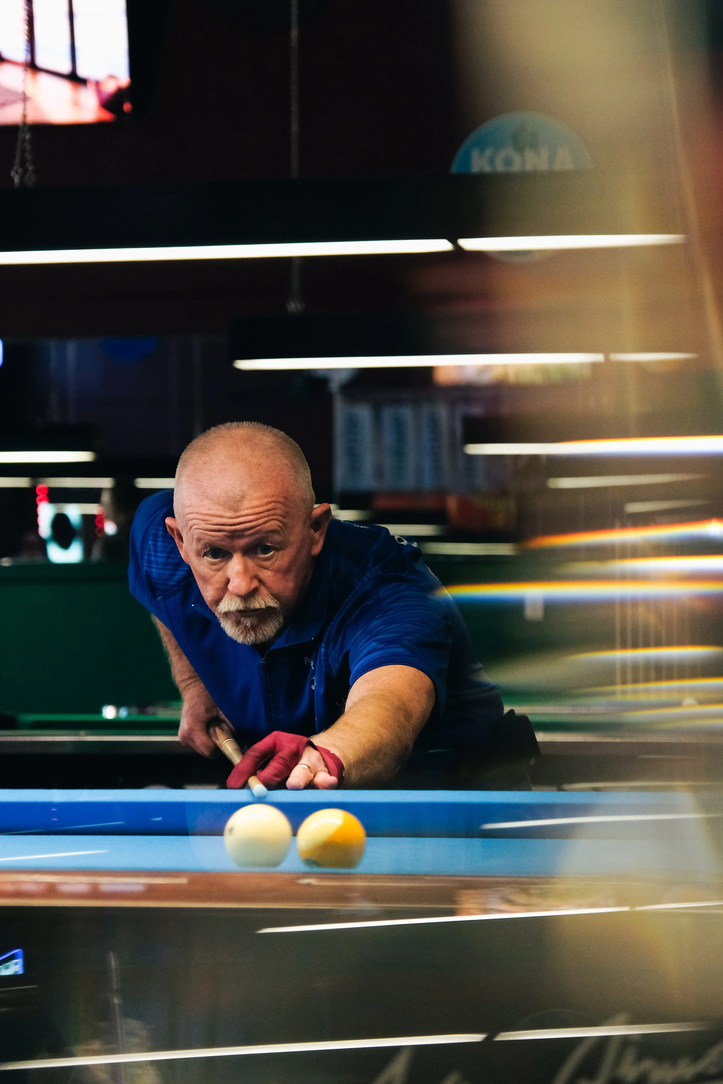 An elderly man with a bald head and white beard playing billiards in a casino or pool hall, focused on a shot, wearing a blue shirt and red glove on his right hand, with balls on the table in front of him.