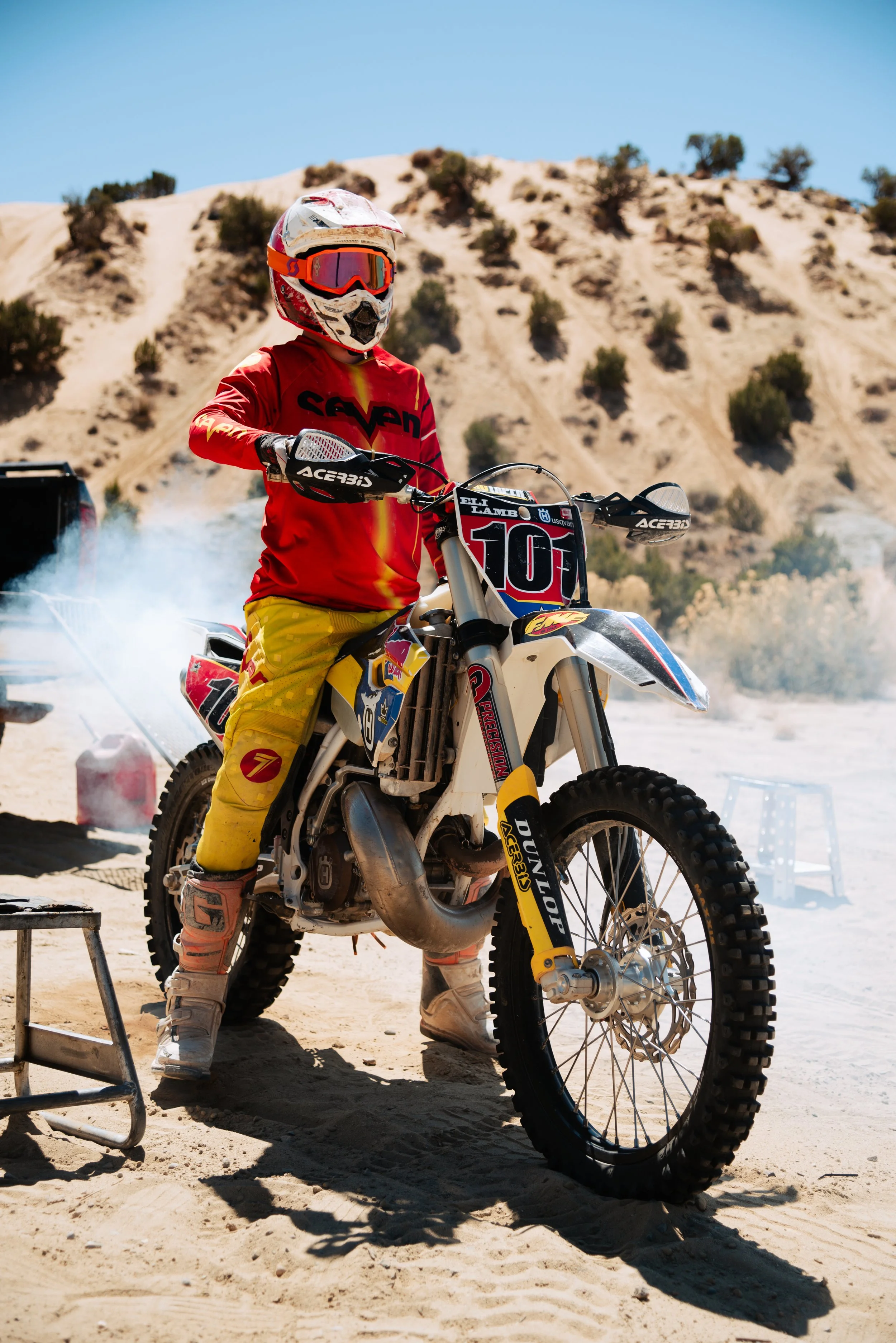 A person wearing a red and yellow motocross outfit with a helmet, goggles, and gloves, standing next to a dirt bike in a desert landscape with hills and sparse vegetation.