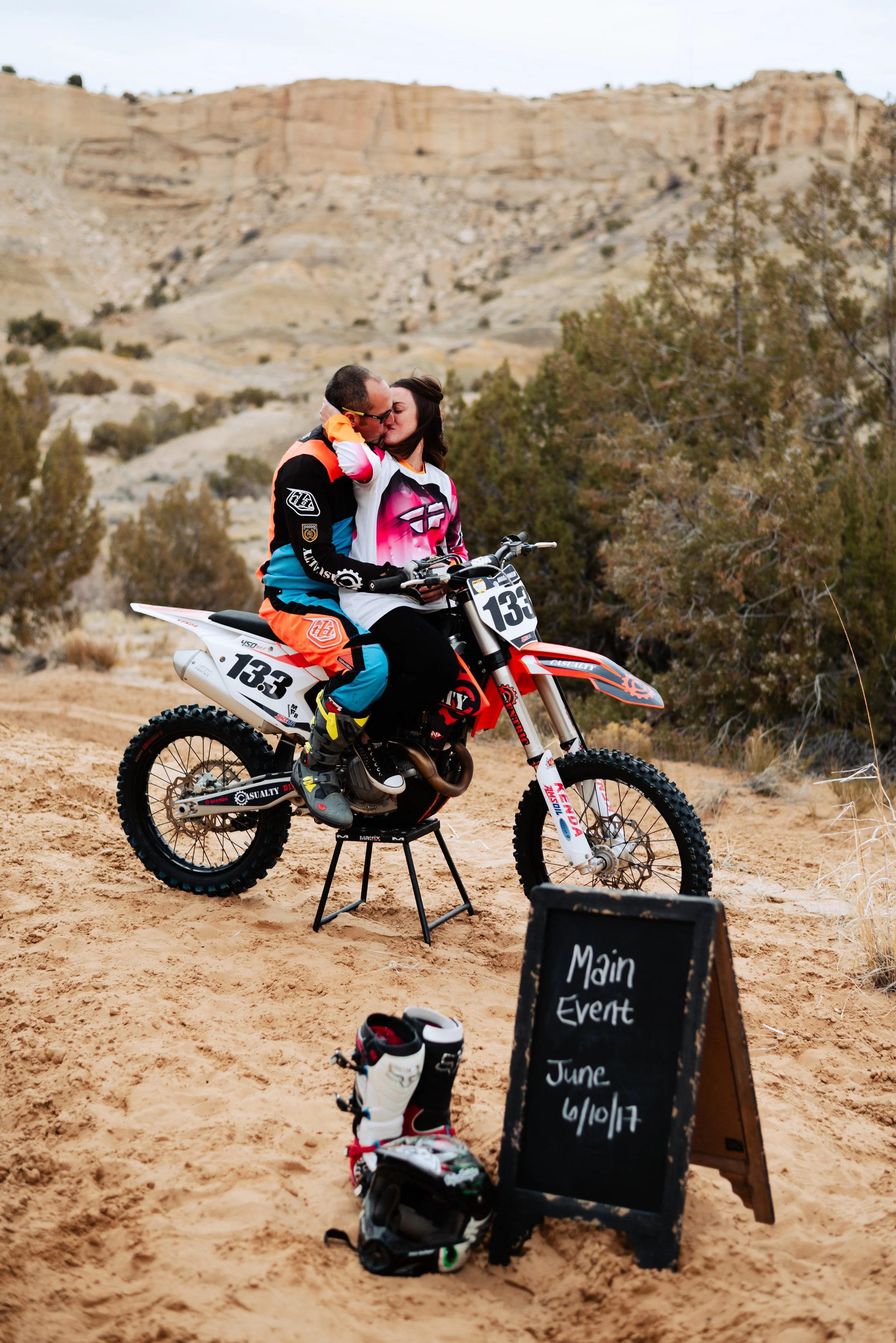 A couple sharing a kiss on a dirt bike in a desert landscape. A blackboard sign with white writing indicates a main event on June 6/10/17. There are motocross gear and boots on the ground.