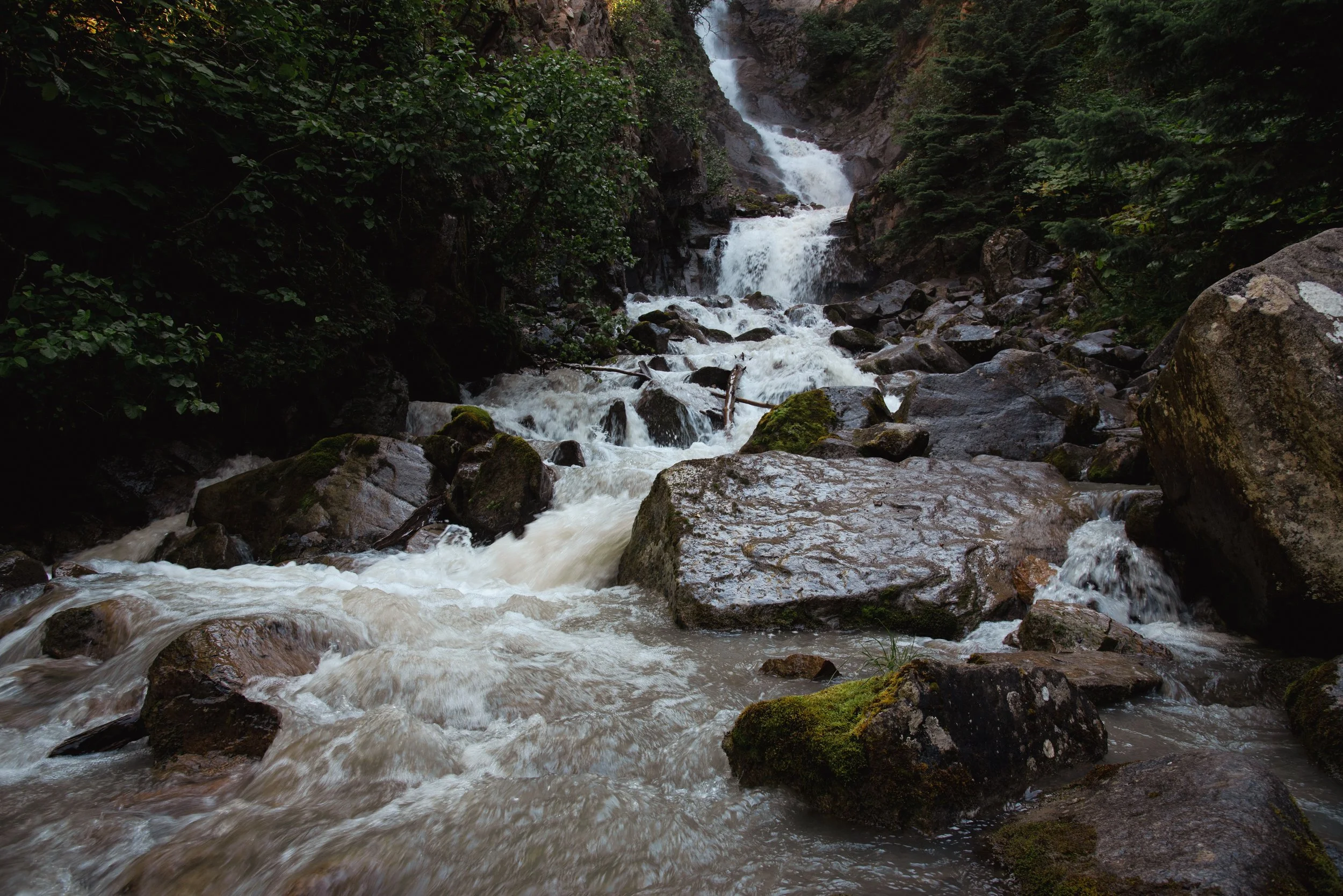 A waterfall flowing through a rocky and forested gorge, with water cascading over moss-covered rocks.