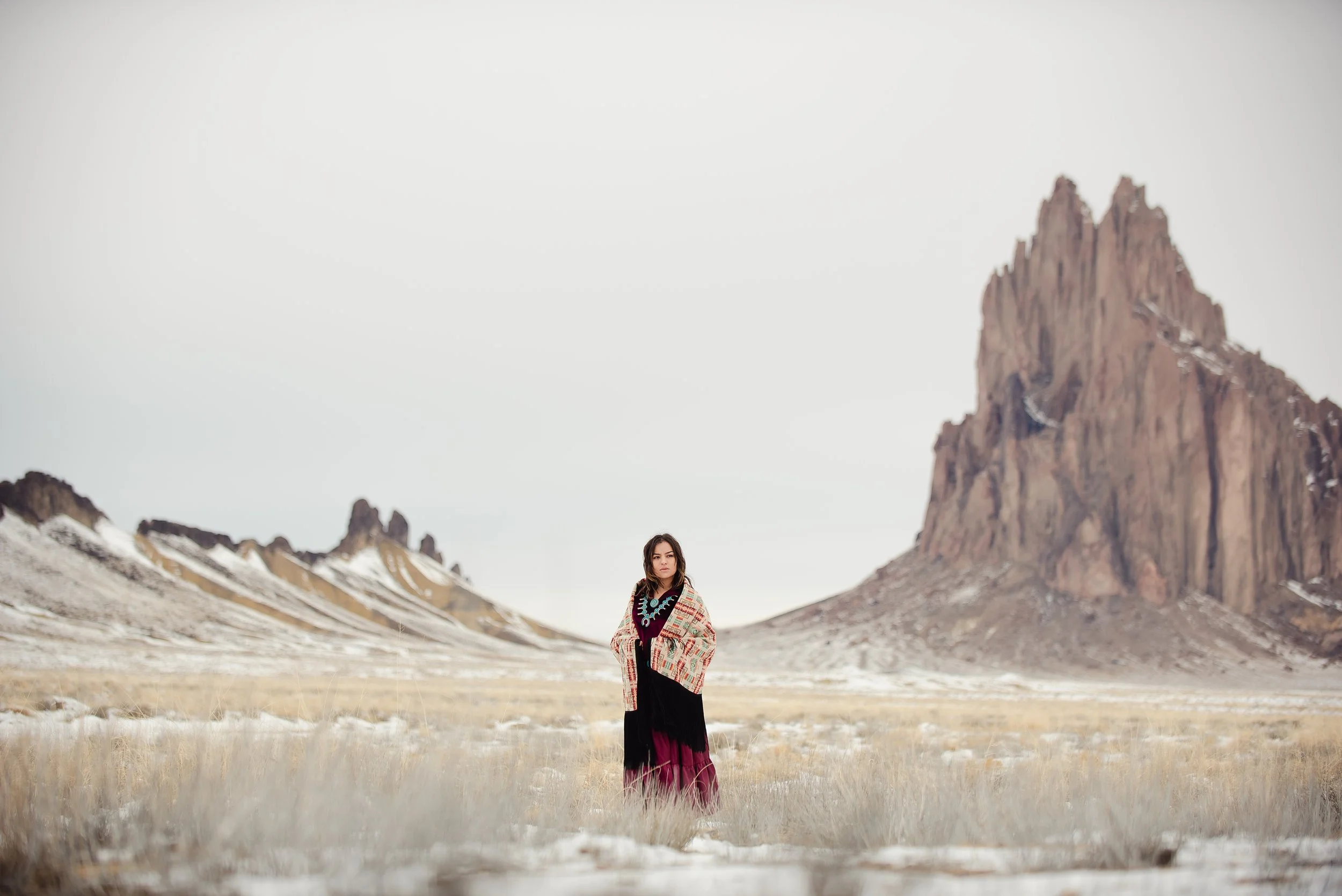 A woman standing in a vast, open desert landscape with rocky mountains in the background, wearing a colorful bohemian-style outfit and a patterned shawl.