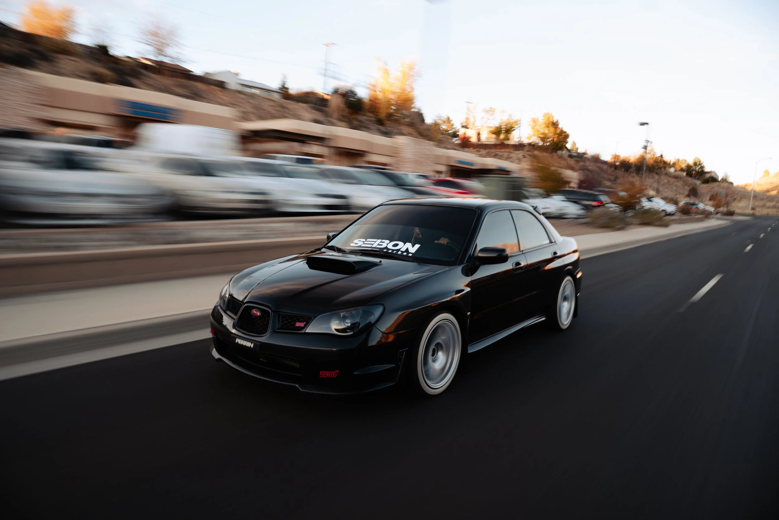 Black sports car driving on a road with a blurred background of parked cars and buildings at sunset.