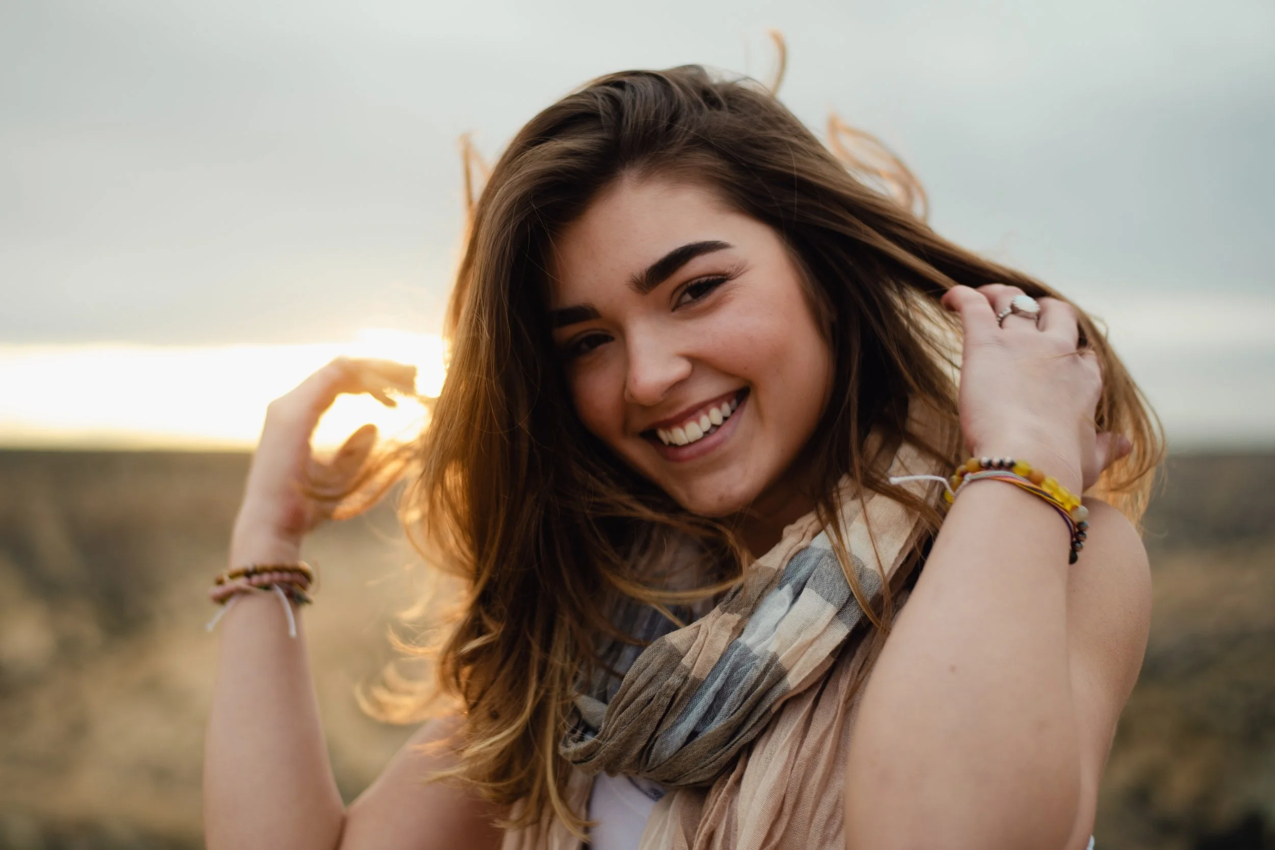A smiling woman with long brown hair, wearing bracelets and a scarf, outdoors at sunset with wind blowing her hair.