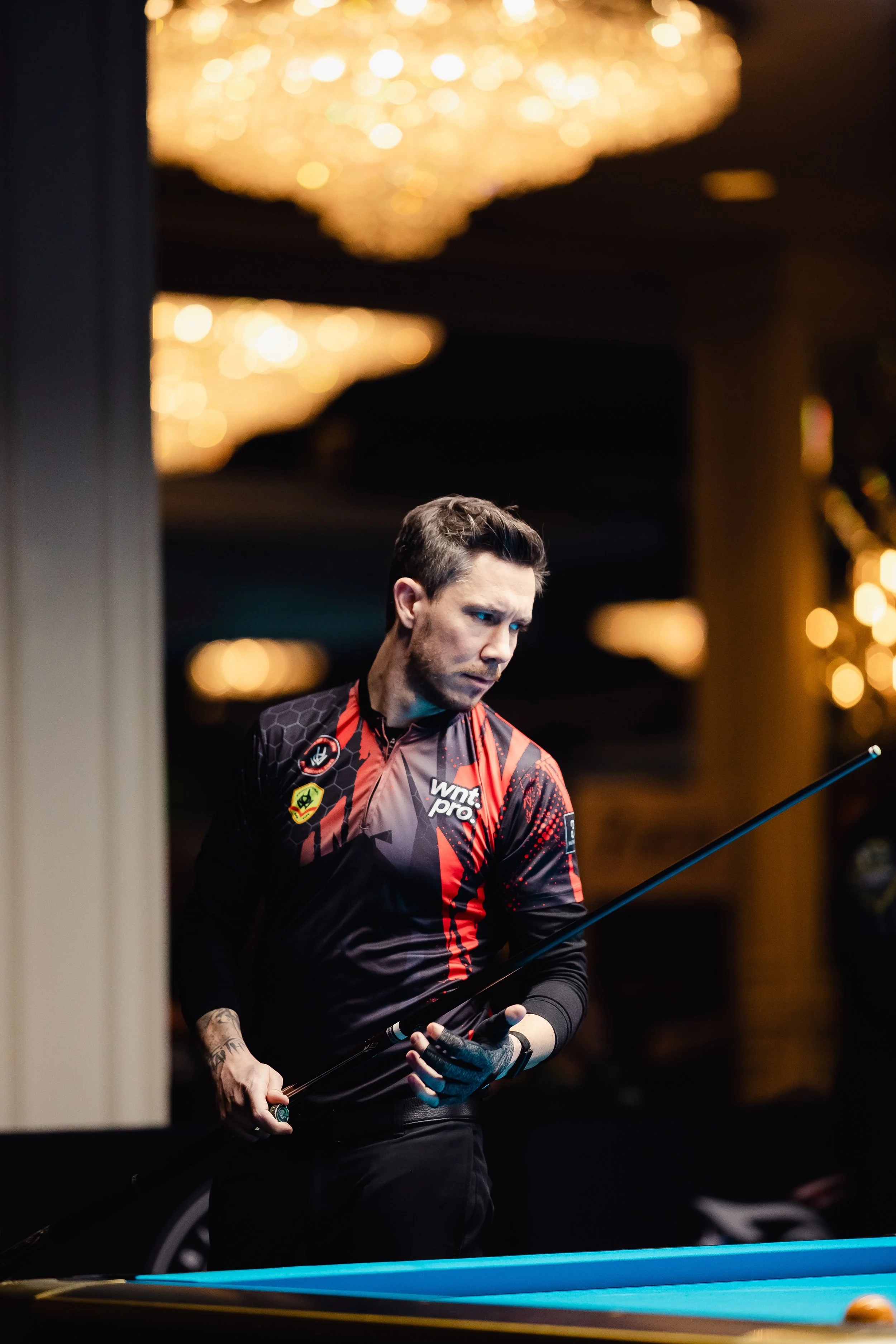 A man in a black and red pool player shirt holding a cue stick, standing near a pool table in a dimly lit room with chandeliers.