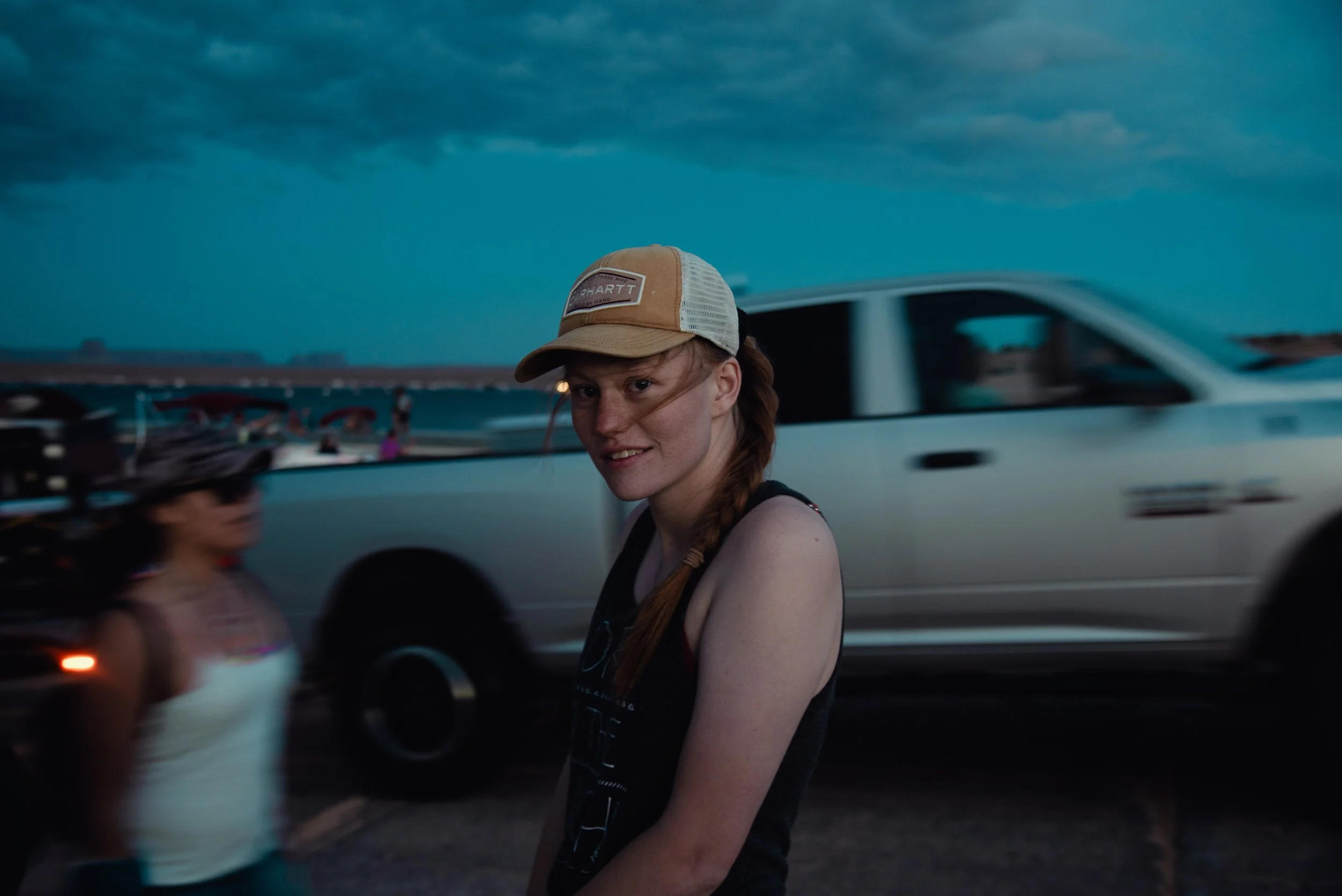 A young woman with red hair and freckles wearing a beige and white trucker cap and a black tank top, standing outdoors under a cloudy sky near a white pickup truck, with another woman in a white swimsuit and a wide-brimmed hat blurred in the backgrou