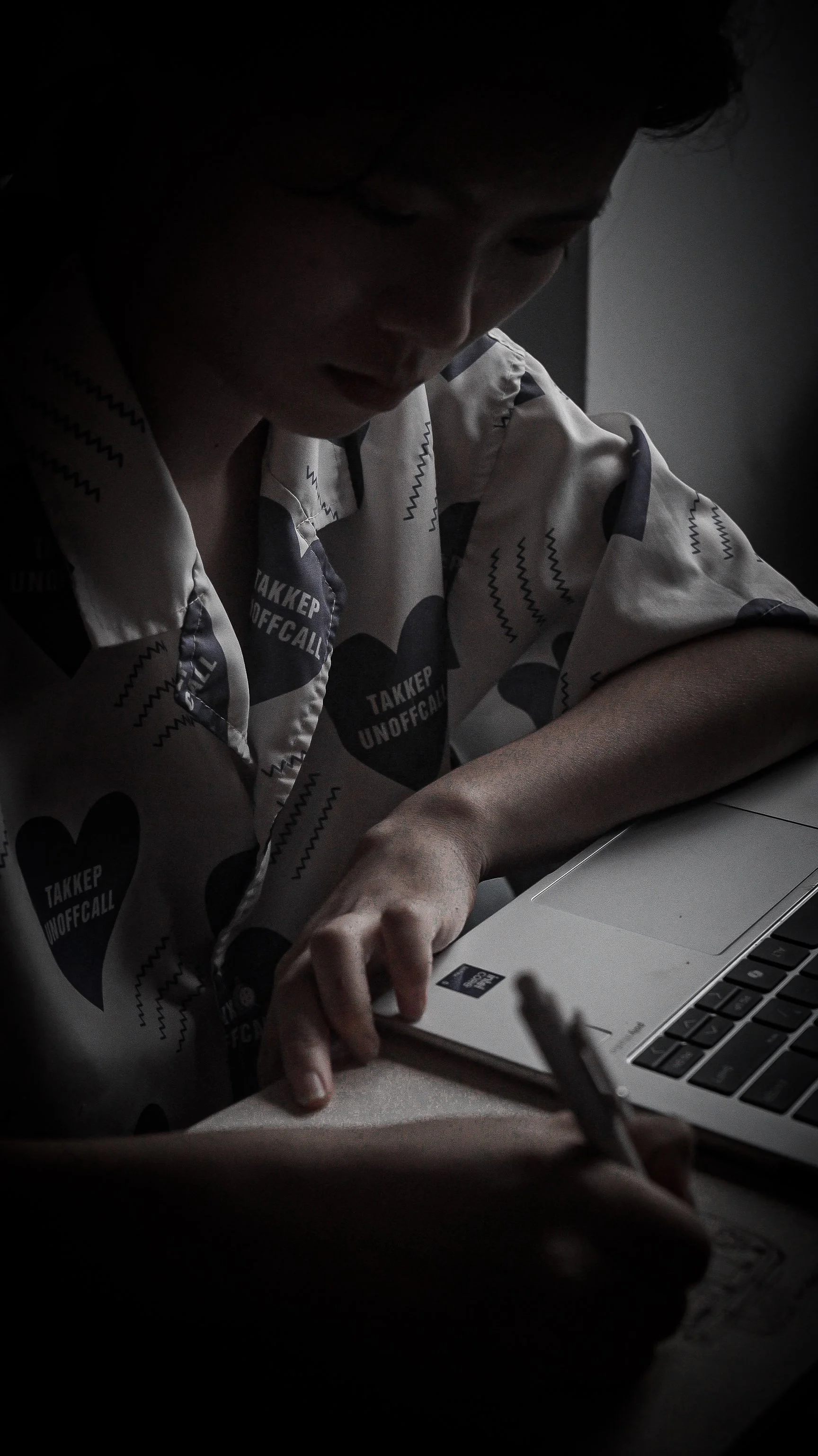 A person wearing a patterned shirt with hearts and the words 'TAKKER UNOFFICIAL' is seen working on a laptop and writing in a notebook.