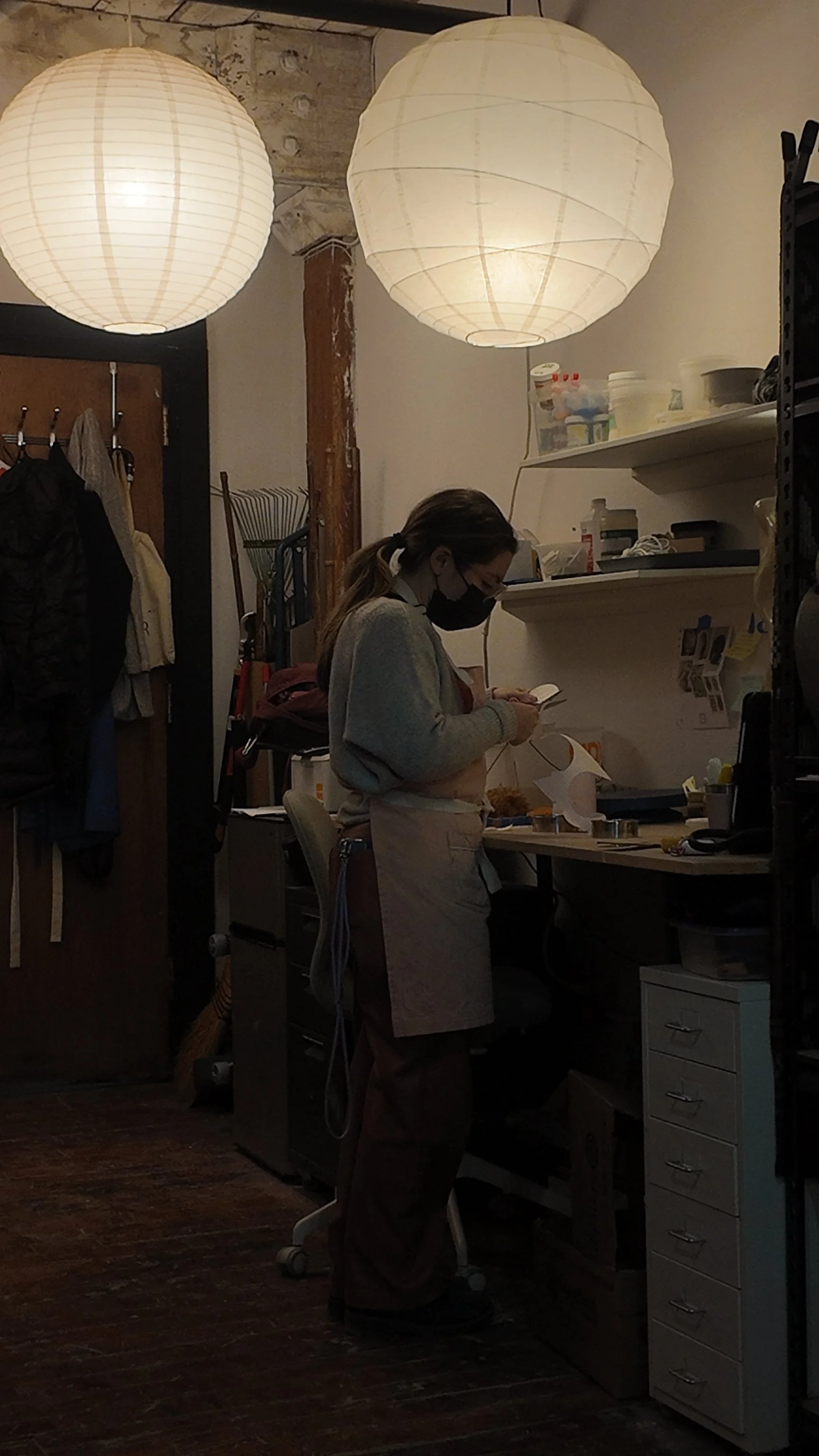 A woman wearing a mask and apron working at a kitchen or workspace station, with shelves, supplies, and hanging clothes around her, illuminated by hanging paper lantern lights.