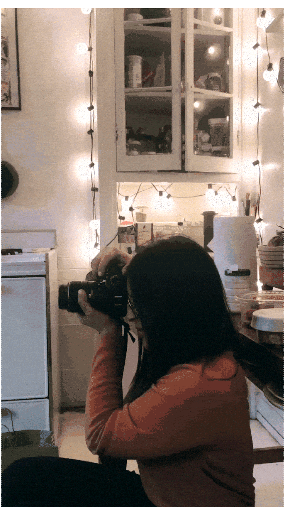 Person taking a photograph in a cozy, decorated kitchen with string lights and shelves.