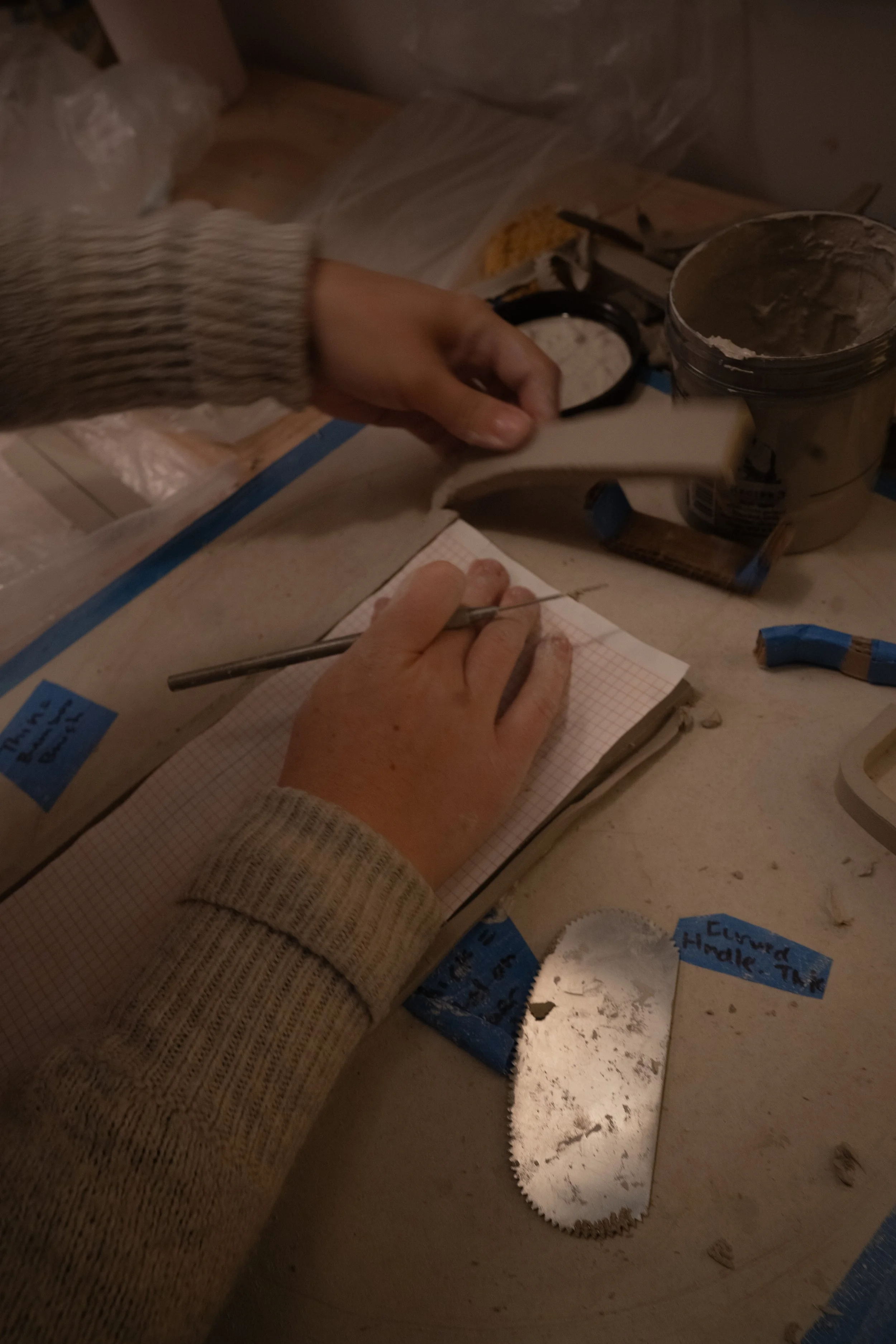 A person working on a craft project at a cluttered workspace, holding a piece of white material with one hand and writing in a notebook with the other. There are tools, a jar of glue, and notes on the desk.