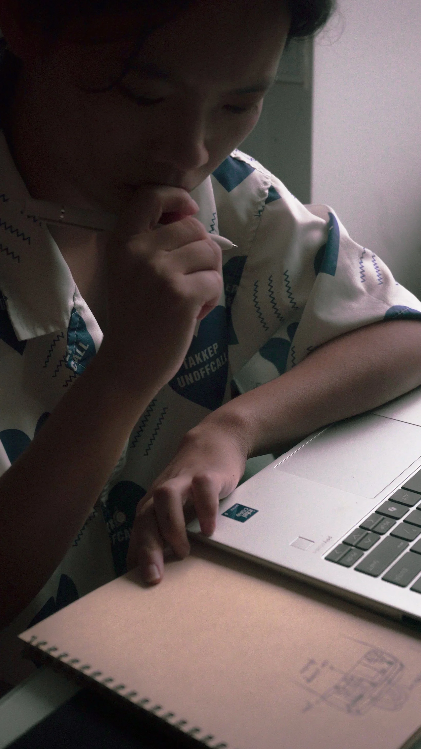 Close-up of a person with dark hair, sitting at a desk, deep in thought, with a laptop and a notebook in front of them.