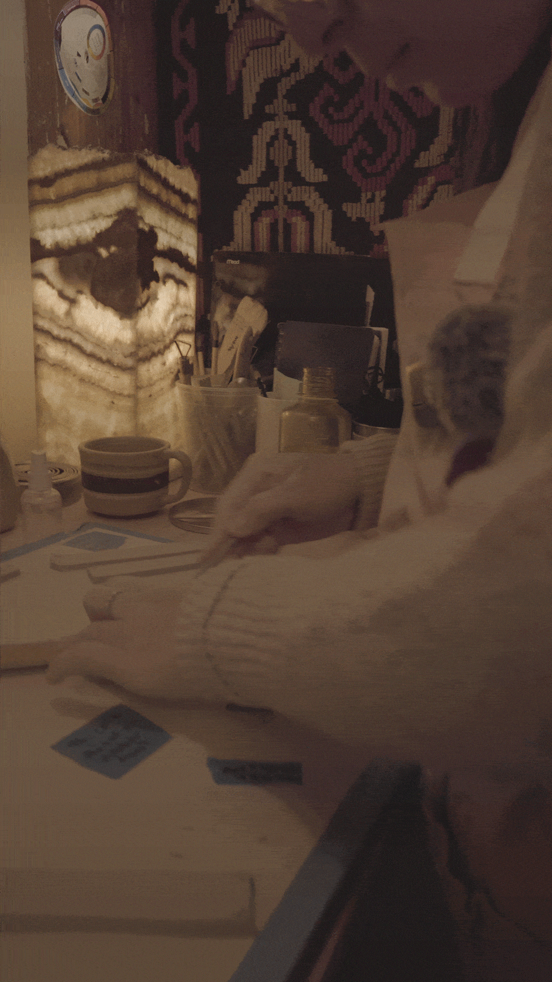A person's arm resting on a table with various items, including cups, clay, and papers, in a dimly lit room with a patterned rug on the floor.