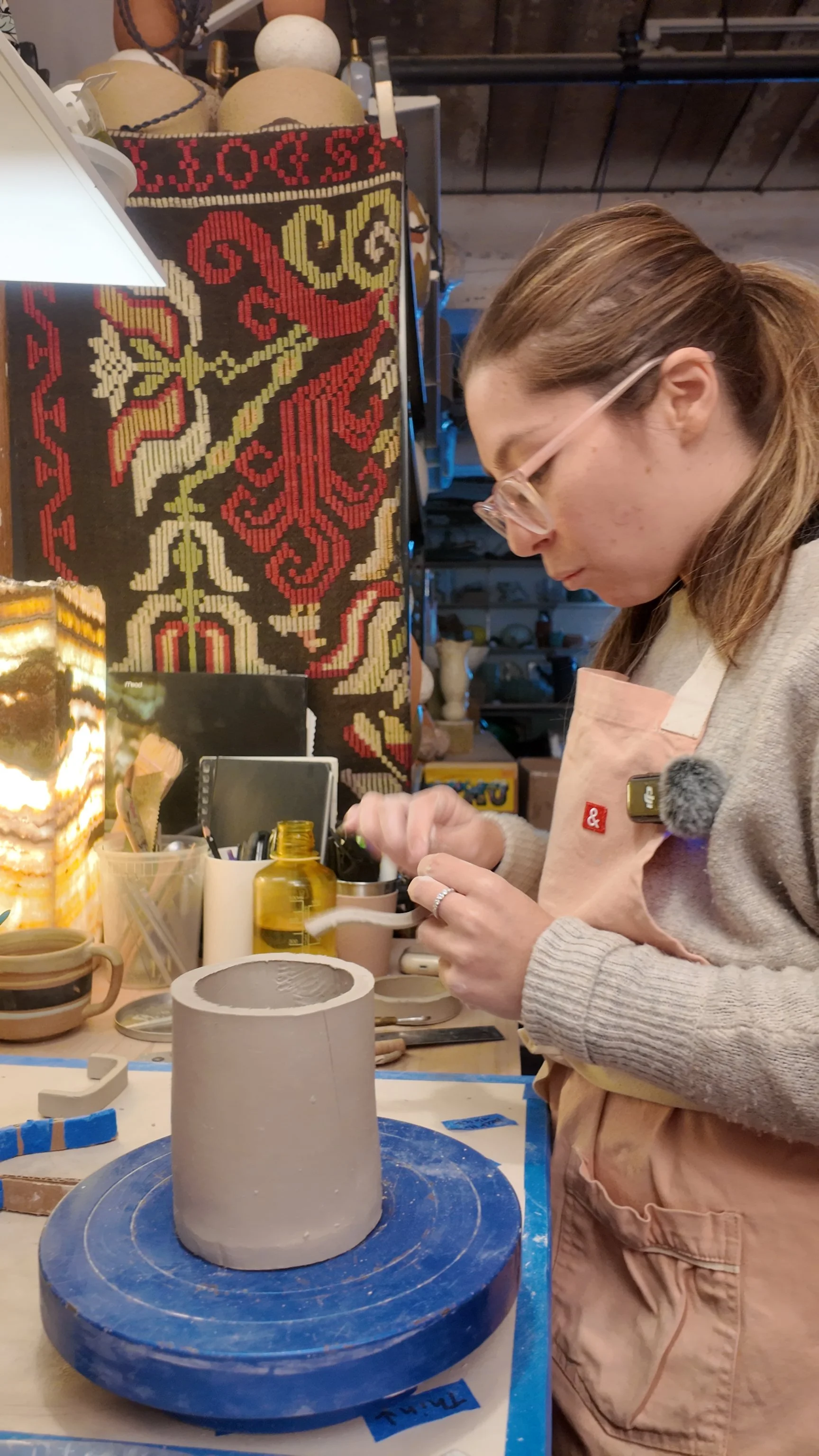 A woman working on a ceramic project in a pottery studio. She is wearing glasses, a beige apron, and a gray sweater. She is shaping or carving a tall, cylindrical ceramic piece on a blue spinning wheel.