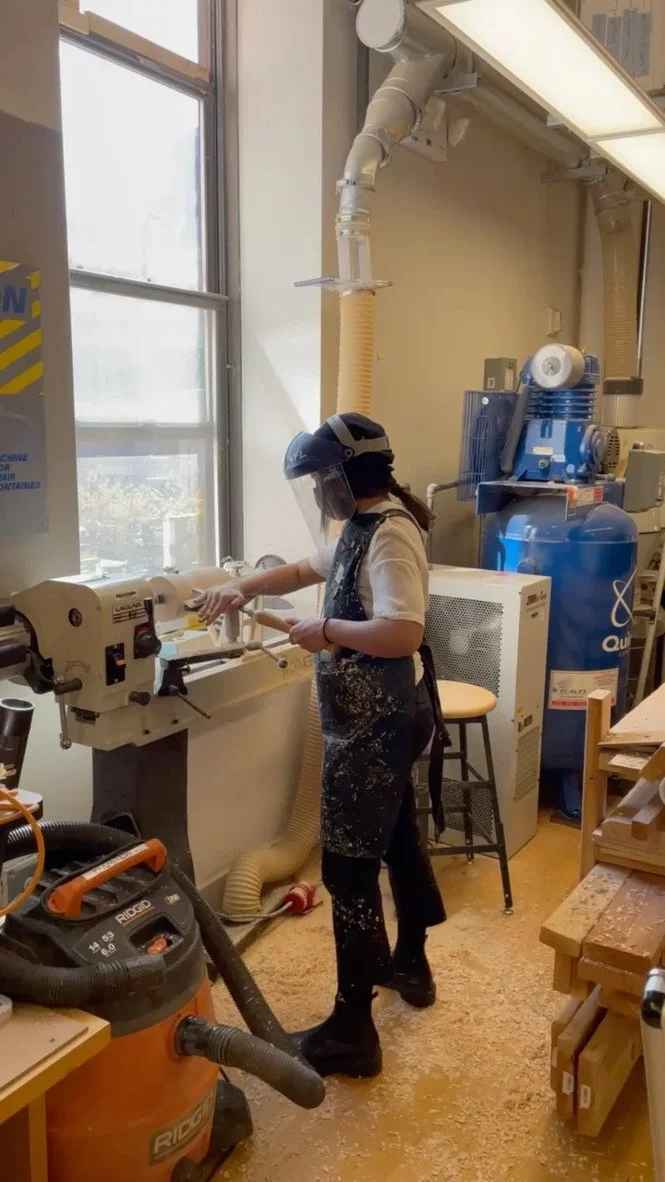 A woman in safety gear, including a face shield and apron, working in a wood workshop with wood shavings on the floor, using woodworking equipment near a large window.
