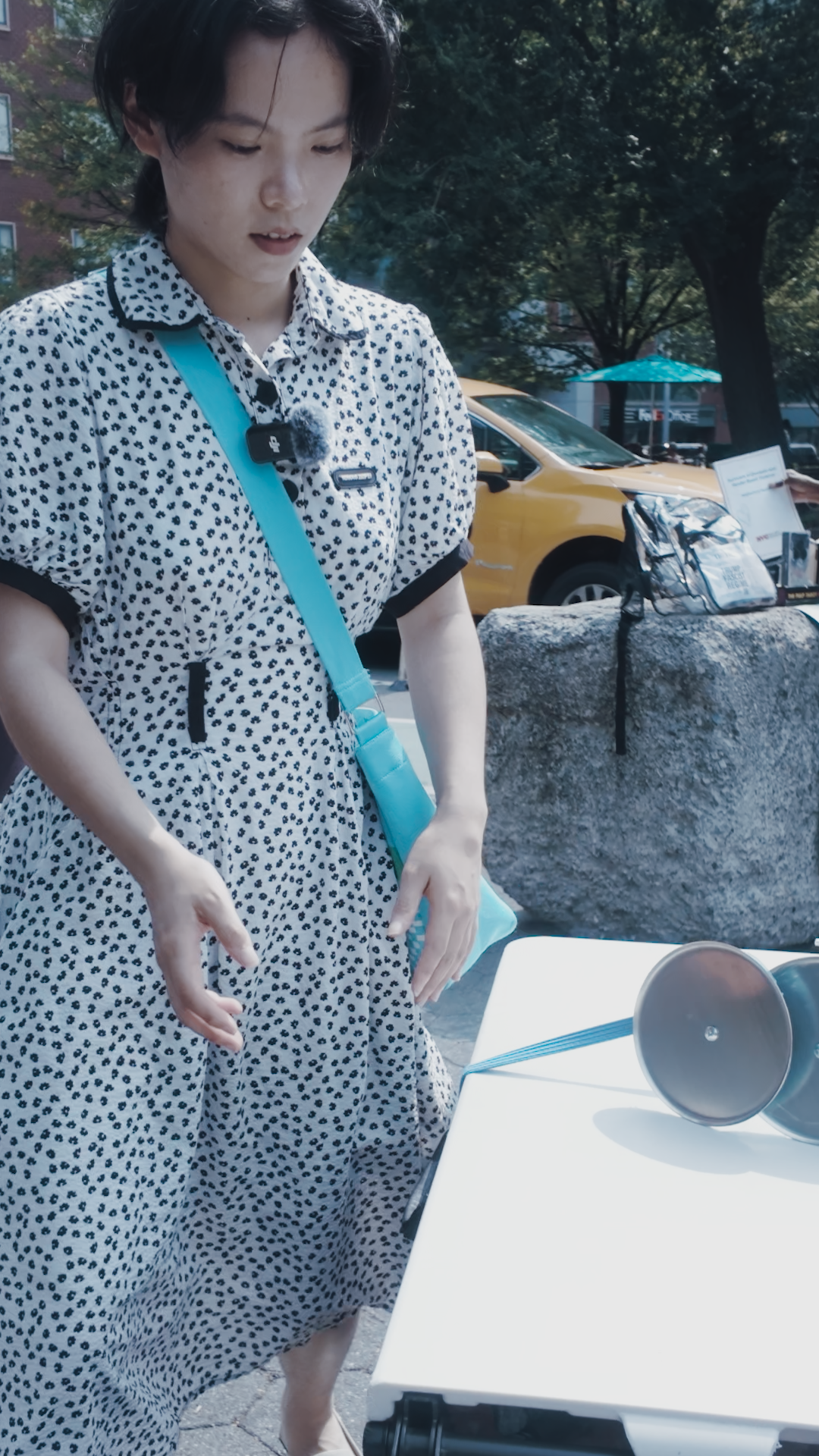 A woman in a polka dot dress standing outdoors next to a white table with a blue megaphone on it. She has a blue crossbody bag and is looking down at the table.