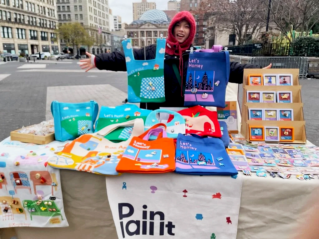 A woman standing behind a table displaying colorful bags, pins, and artwork at an outdoor market, with her arms outstretched and smiling, in an urban setting in the cold.