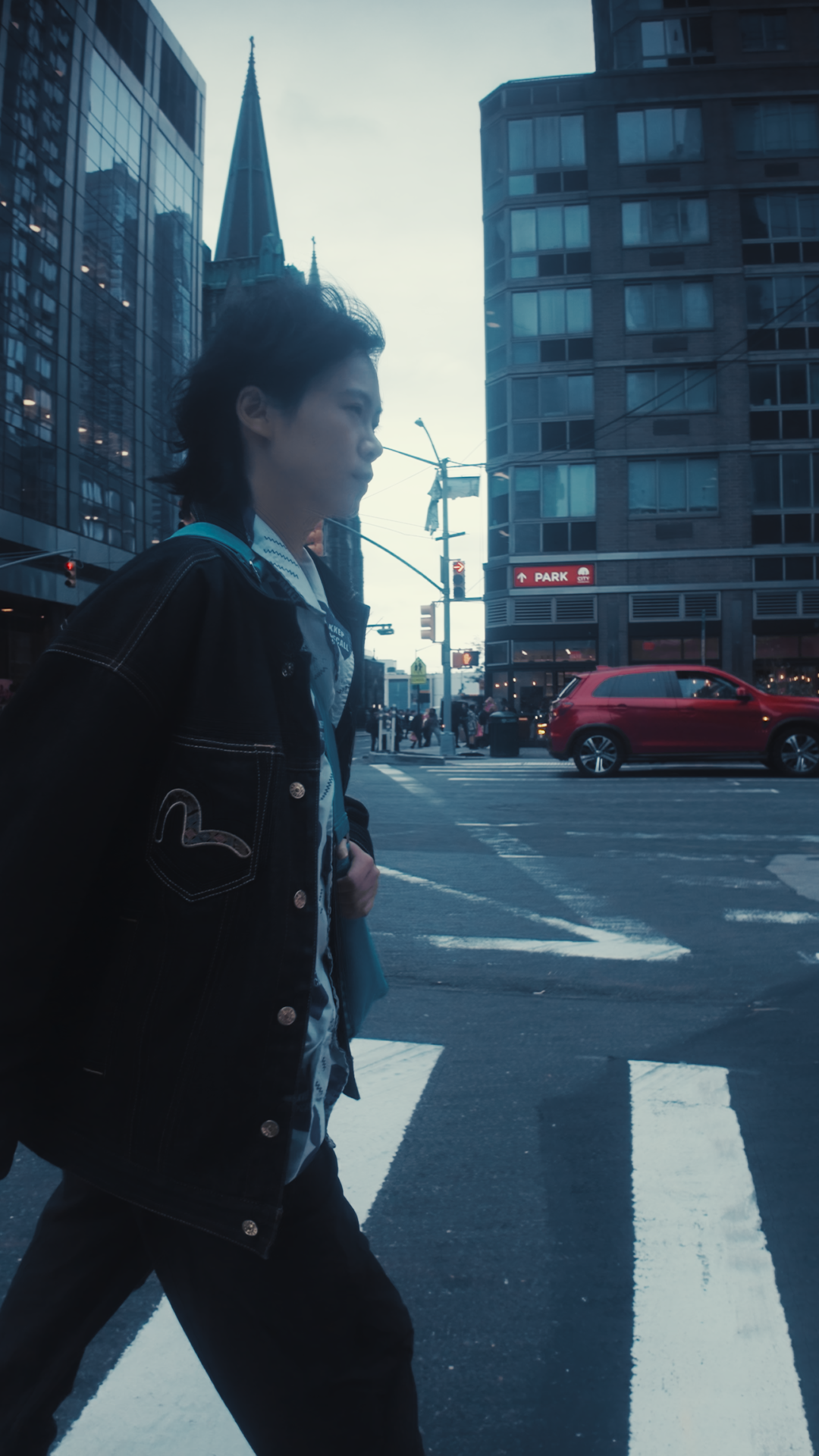 A person with dark hair walking across a city crosswalk with tall buildings, a red car, and a street sign that says 'Park' in the background.
