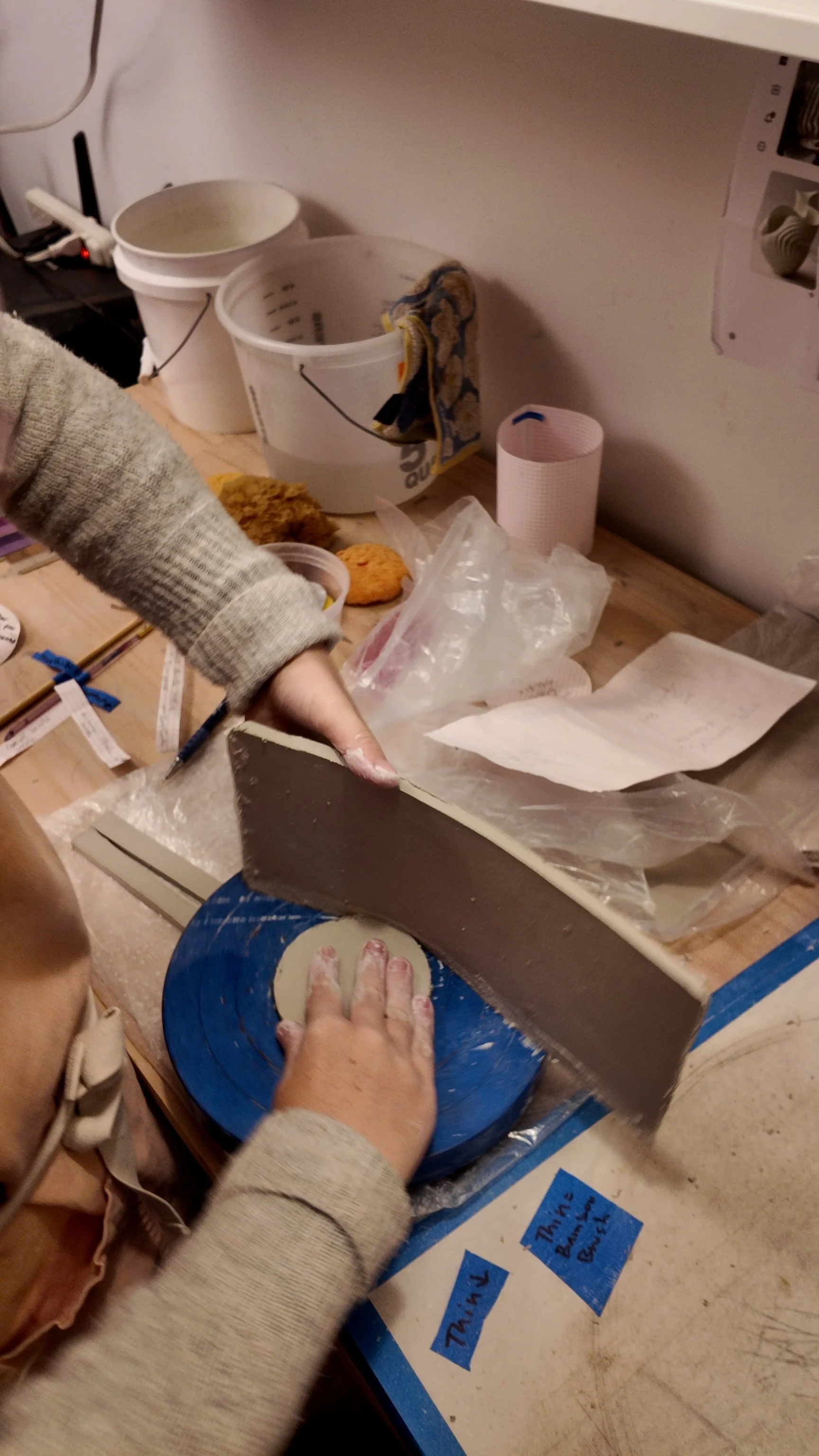 Person cutting clay with a large knife on a blue cutting board in a workshop setting, with various other items on the countertop.