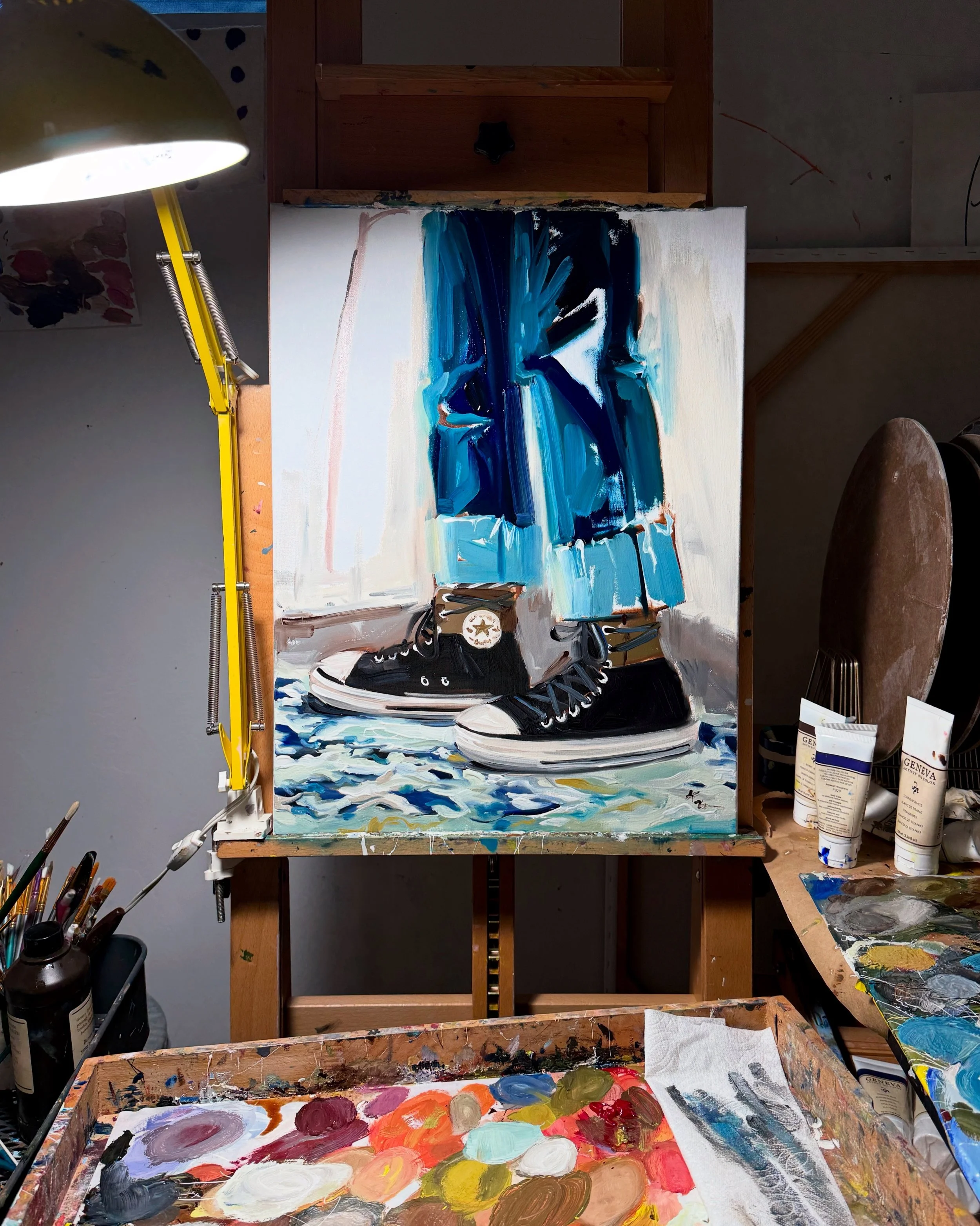 An oil painting in progress of a pair of black sneakers with white soles and blue jeans, standing on a textured surface with splashes of blue and white paint.