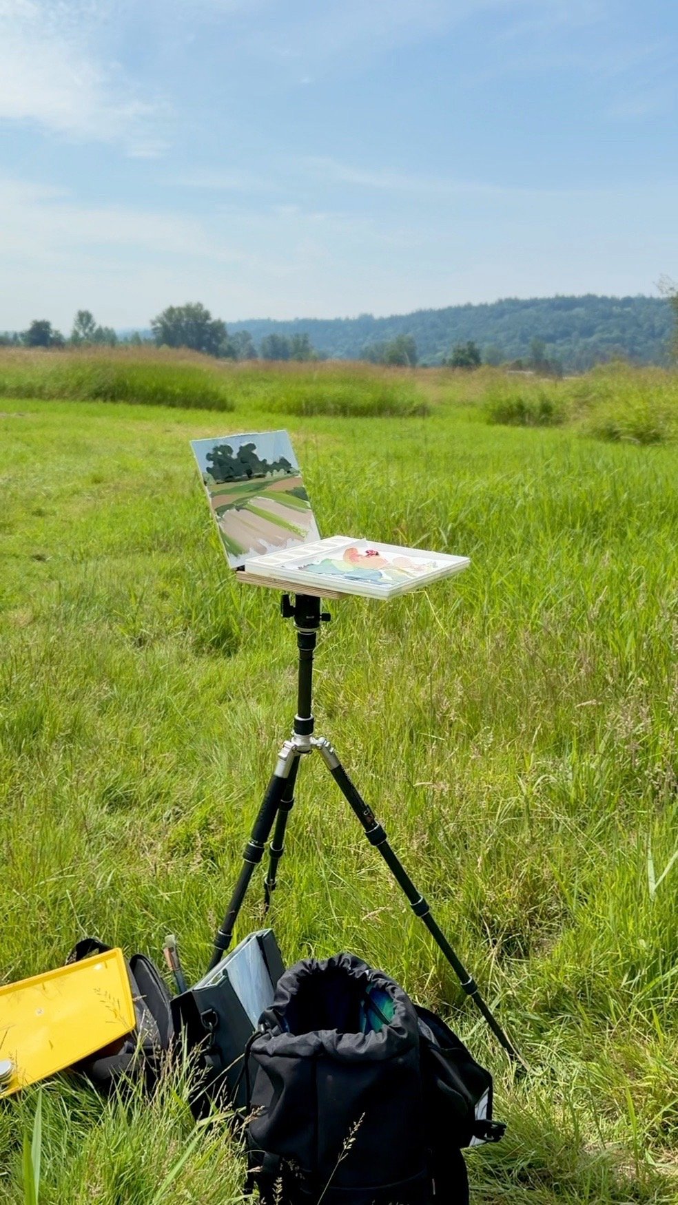 An outdoor scene with a portable easel or painting setup on a tripod in a grassy field. The easel holds a canvas with painted landscape, and there are art supplies, a backpack, and a yellow pencil case on the ground nearby. The background features ro
