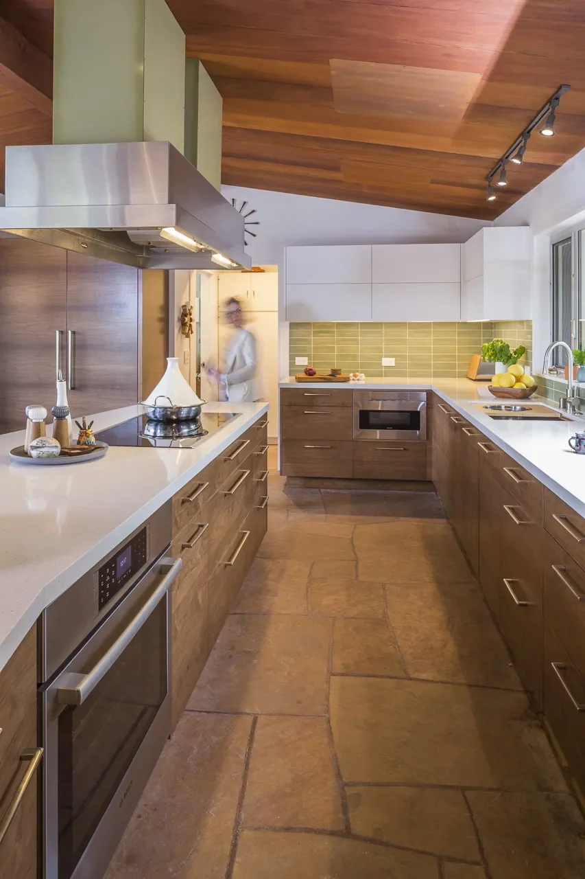 Interior designer Jodi Eisner standing in a Topanga Canyon organic mid-century modern kitchen featuring walnut cabinetry, green tile backsplash, quartz waterfall island, and original stainless steel range hood.