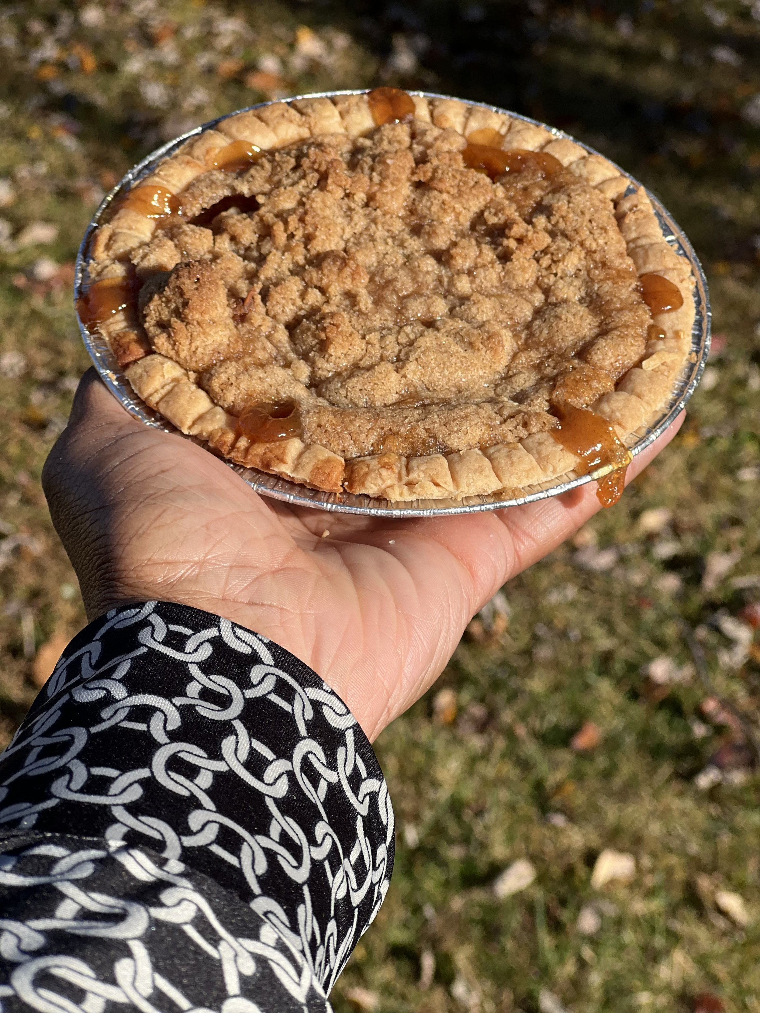 Close-up of a hand holding a peach cobbler pie in a foil container outdoors on grass, with caramel drizzle around the edge.