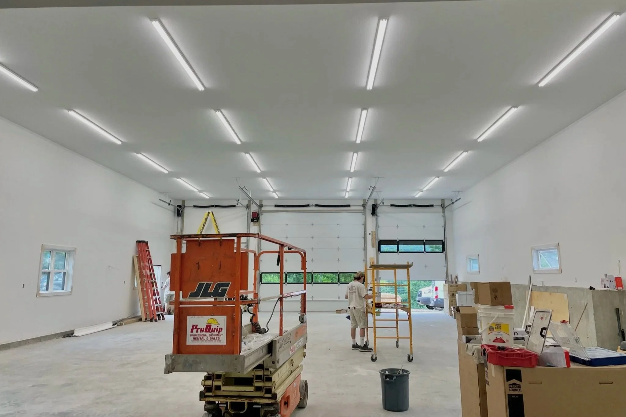 Interior of a large industrial garage under construction with white walls, a high ceiling with multiple fluorescent lights, and small windows. Construction tools and equipment, including a scissor lift, ladder, and various boxes and materials, are sc