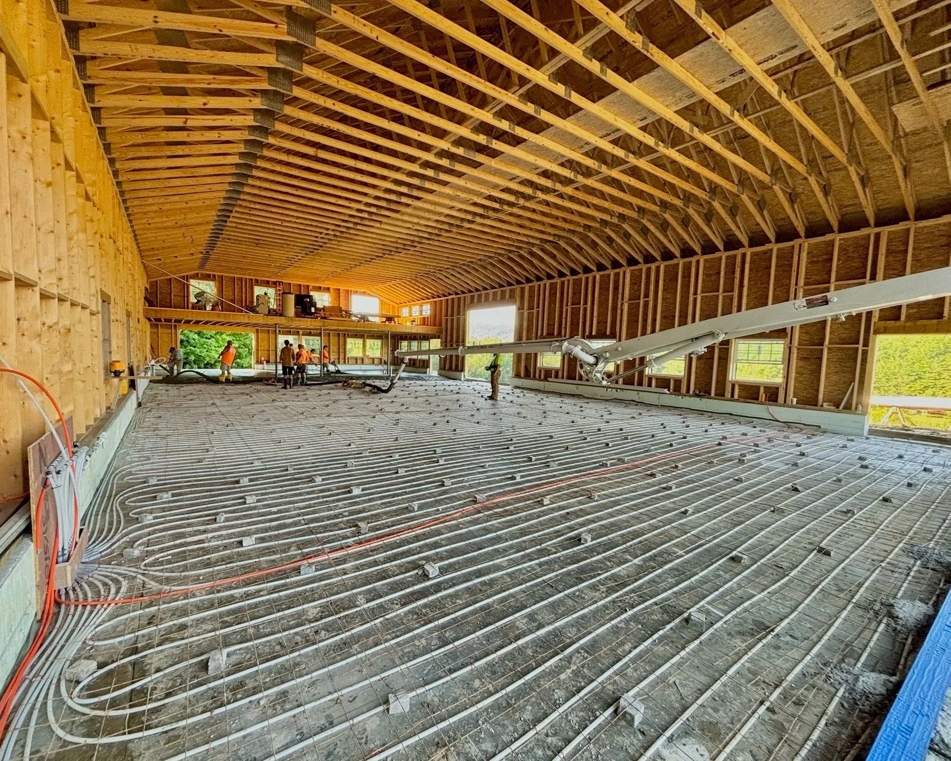 Interior view of a construction site with workers installing radiant floor heating system in a large building, with wooden framing and multiple window openings.