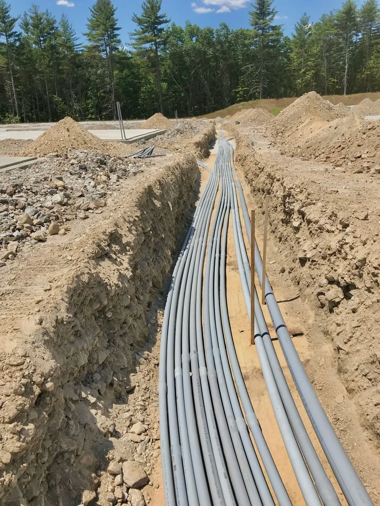 Construction site with electrical conduits laid in a trench, surrounded by dirt and gravel, with trees in the background.